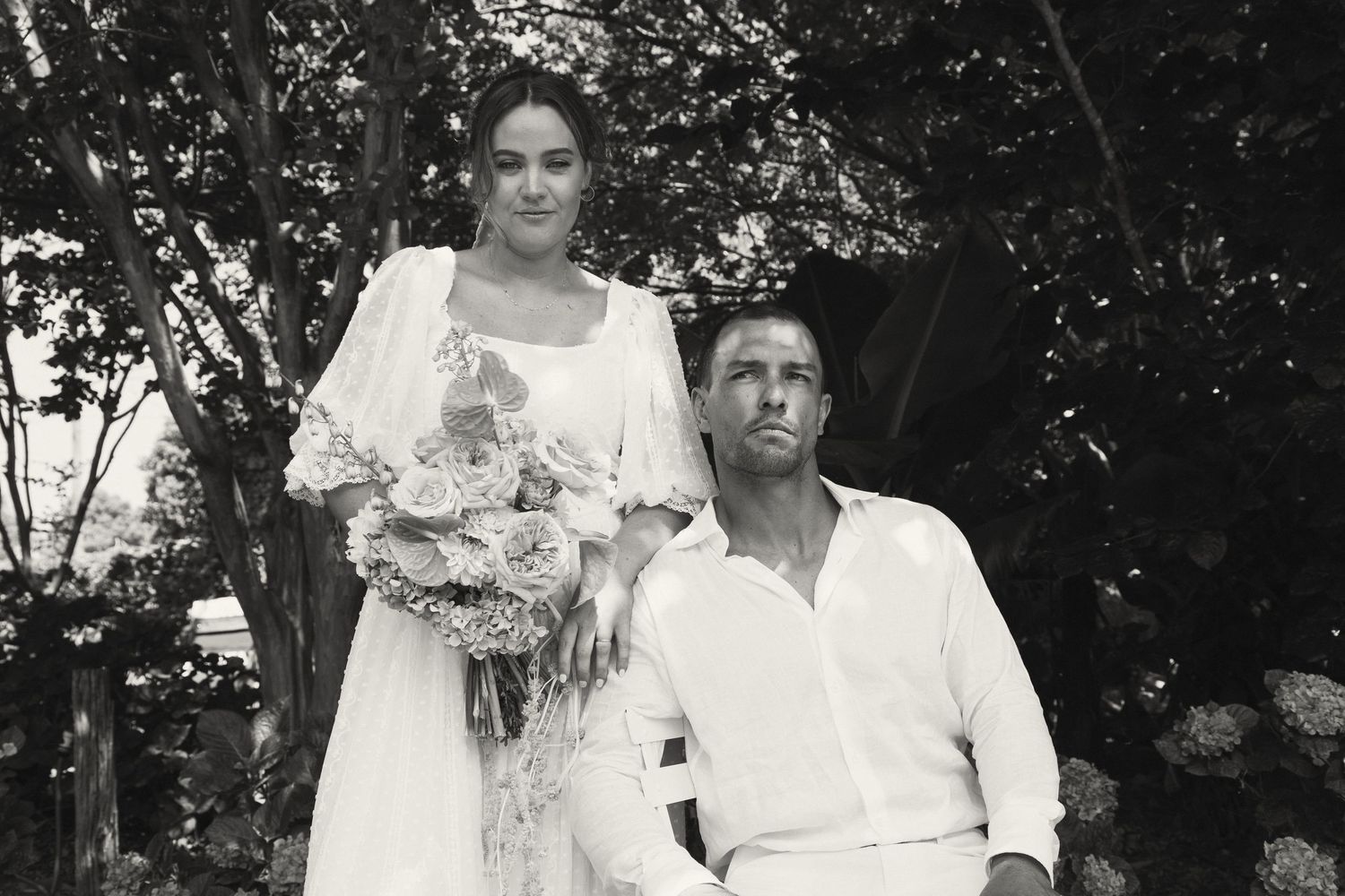 A vintage black and white portrait of a couple outdoors with the bride holding flowers and wearing a white dress.