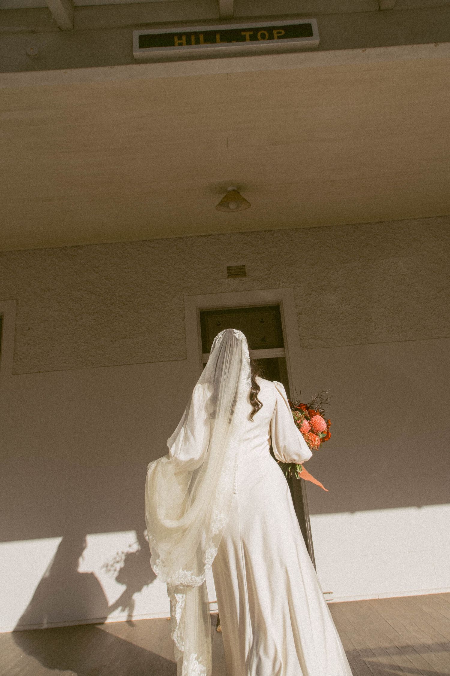 A bride in a long white dress and veil enters a building entrance.