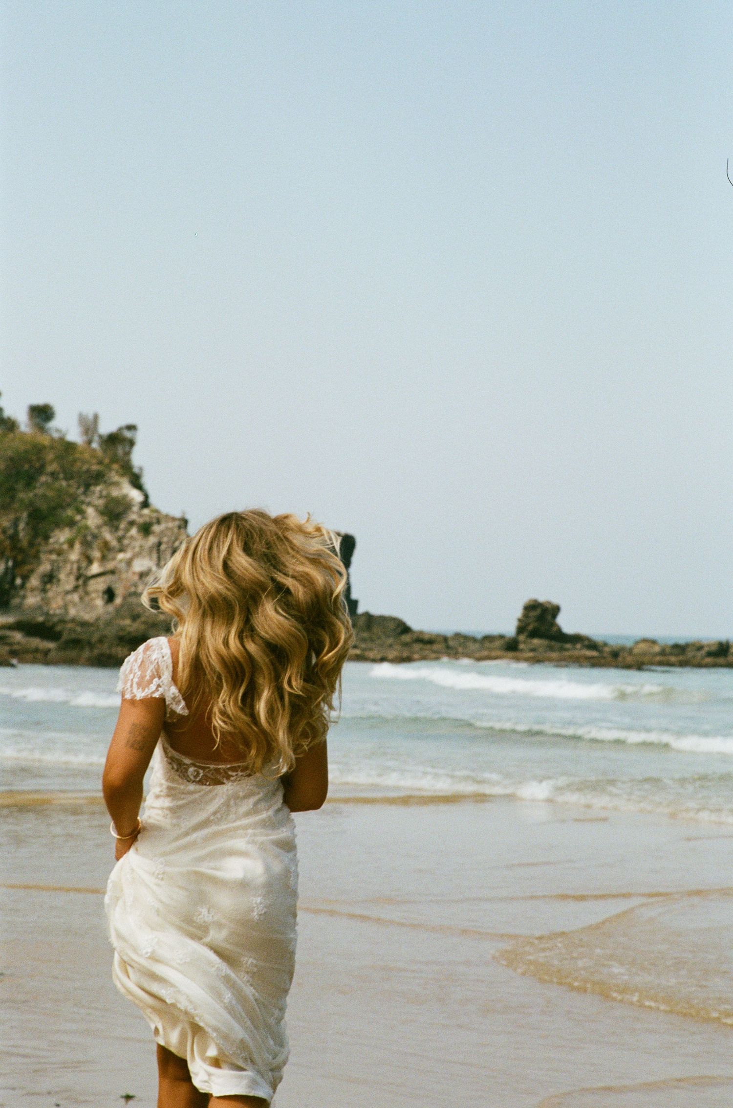 A white dress flows in the ocean breeze as someone walks along a scenic beach with rocky cliffs in the background.