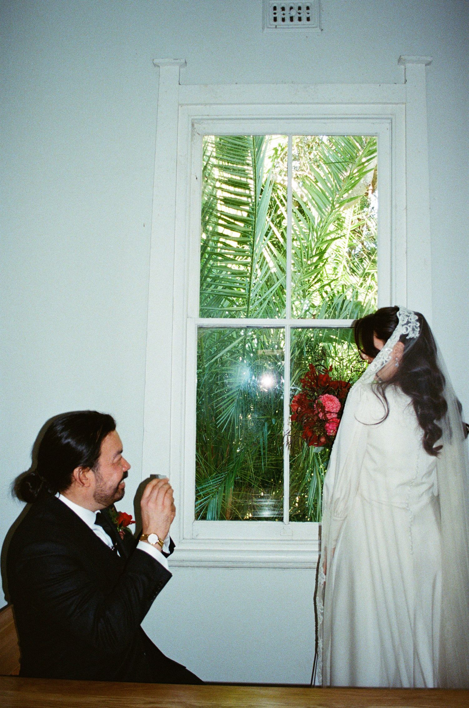 A playful wedding scene by a window with palm tree views as two pose in formal attire.