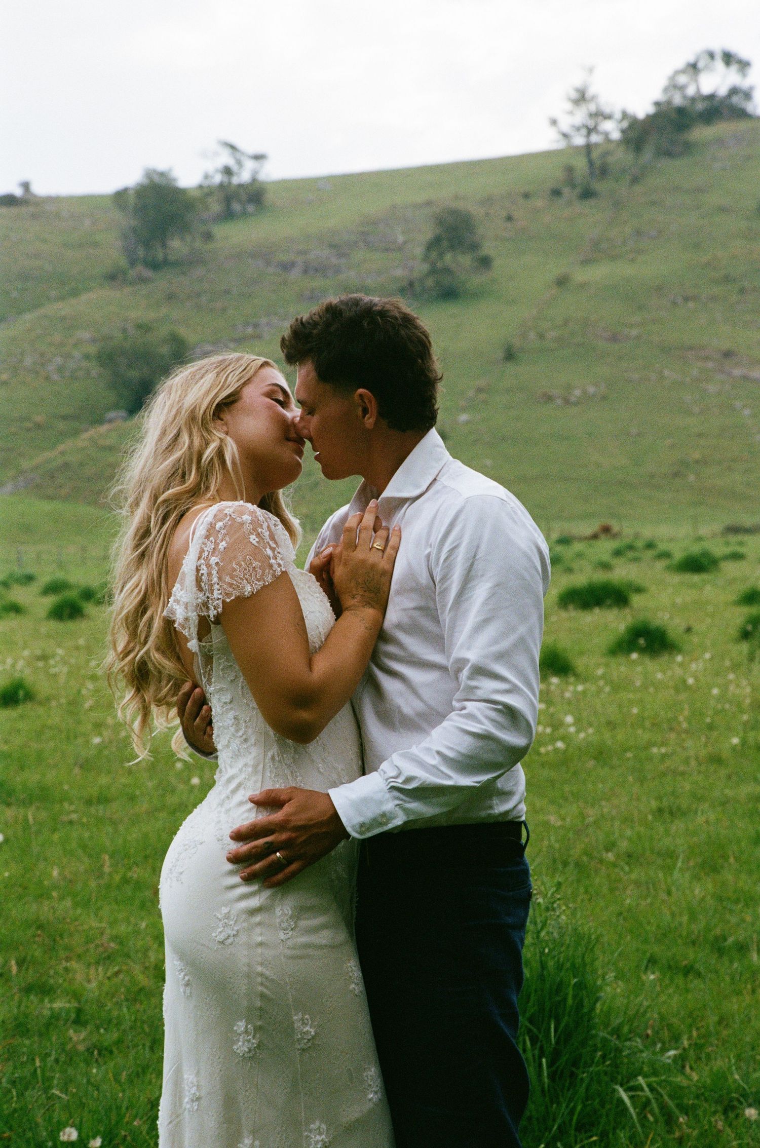 A romantic wedding couple shares intimate kisses against a scenic green hillside backdrop.
