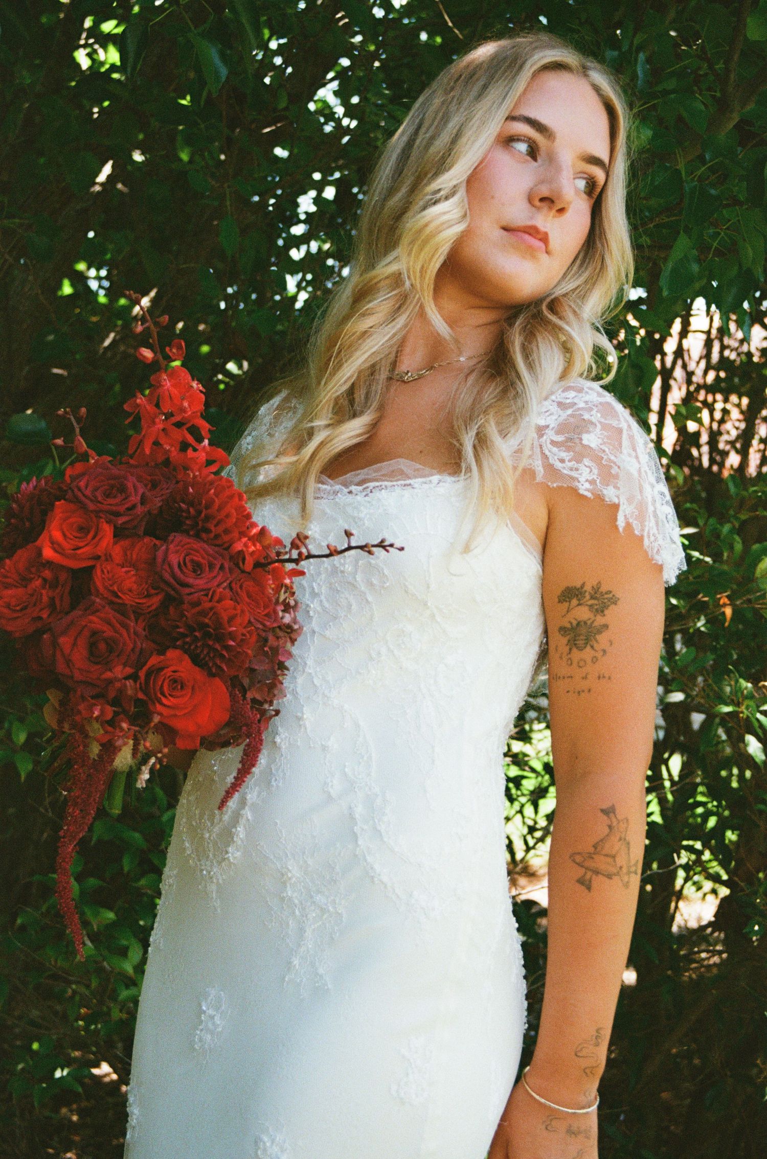 A bride in a lace wedding dress holds a vibrant bouquet of red roses against a backdrop of green foliage.