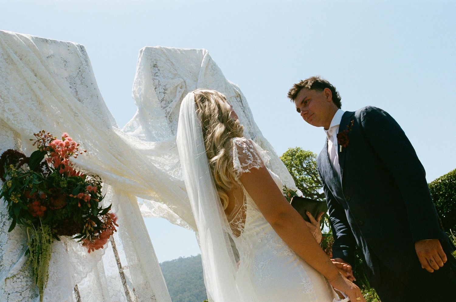 A bride and groom share a romantic moment outdoors with flowing veil and red floral bouquet on a sunny day.