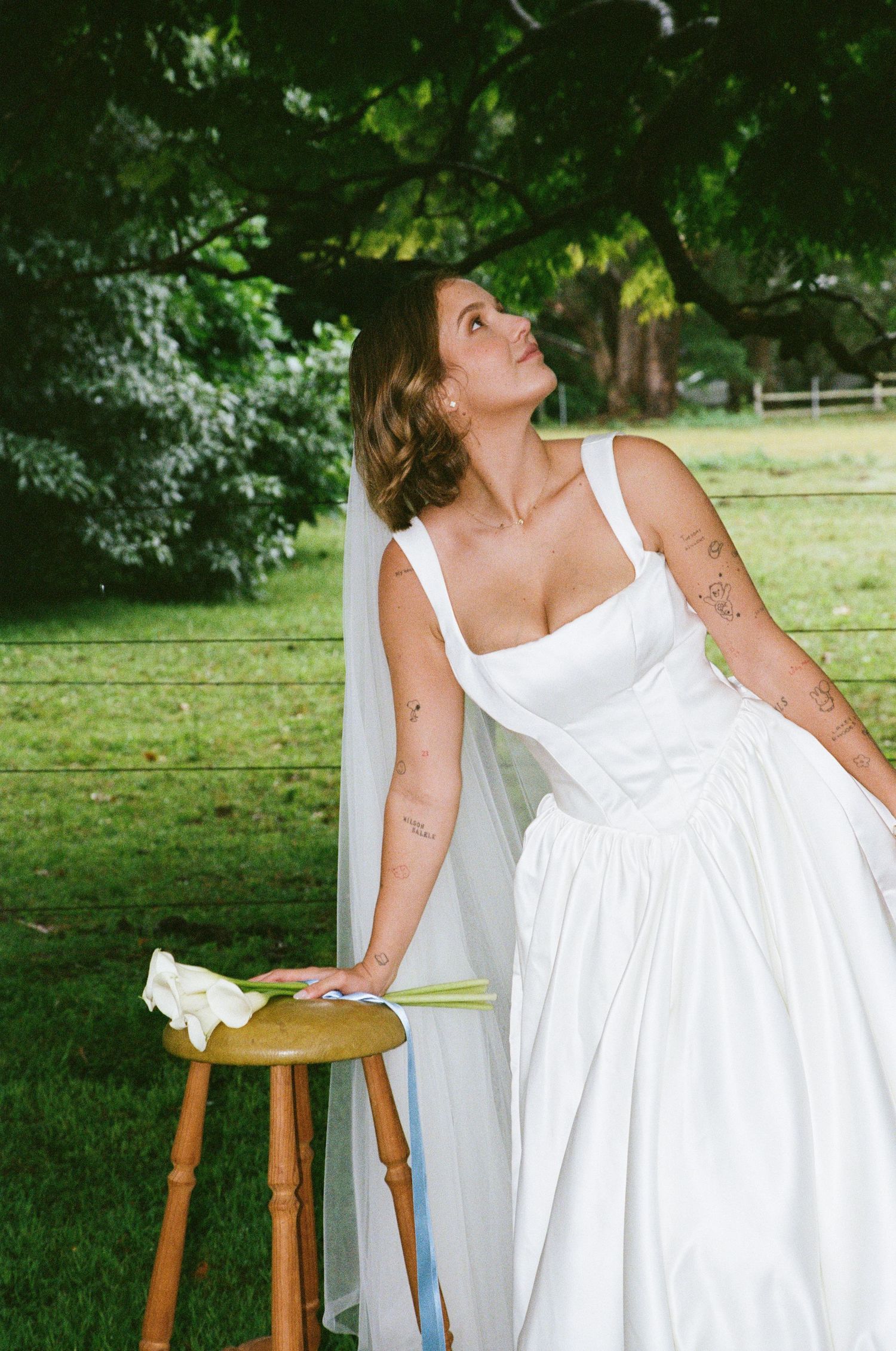 A flowing white wedding dress is artfully displayed against a lush green garden backdrop with a wooden chair.