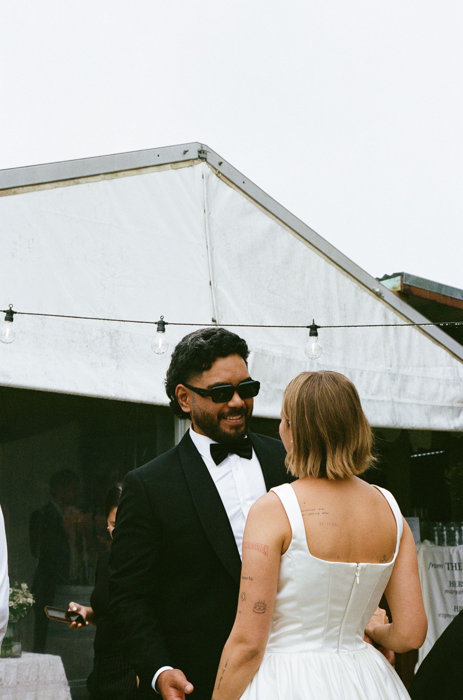 A couple shares a moment under a white tent at a formal outdoor event.