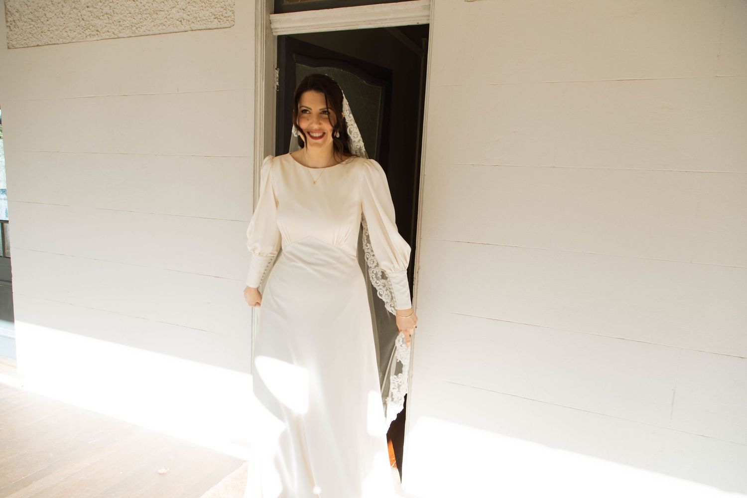Someone stands in a doorway wearing a vintage-inspired cream-colored dress with long sleeves and elegant drapery.