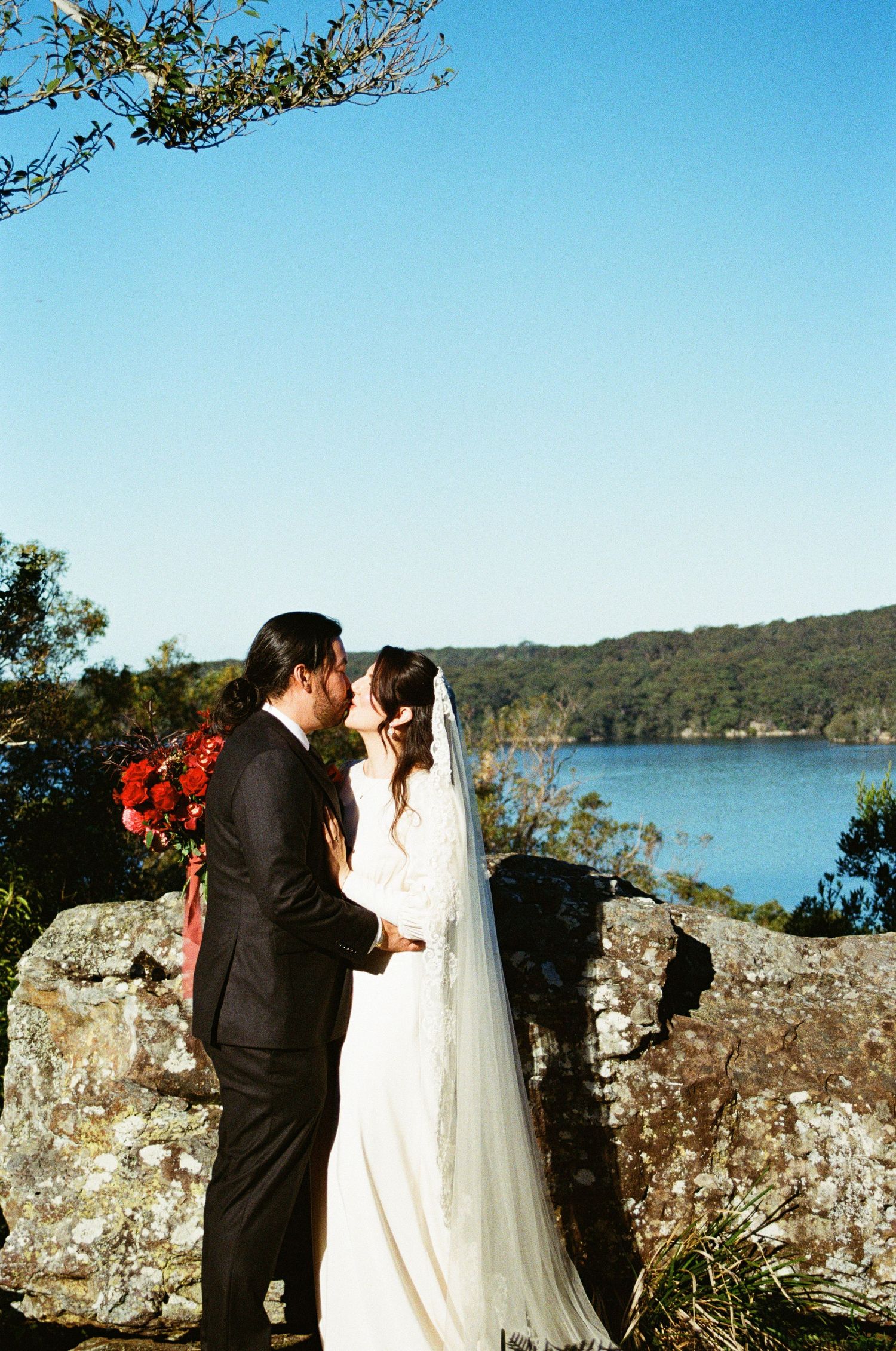 A couple in wedding attire standing on rocky cliffs overlooking a scenic lake.