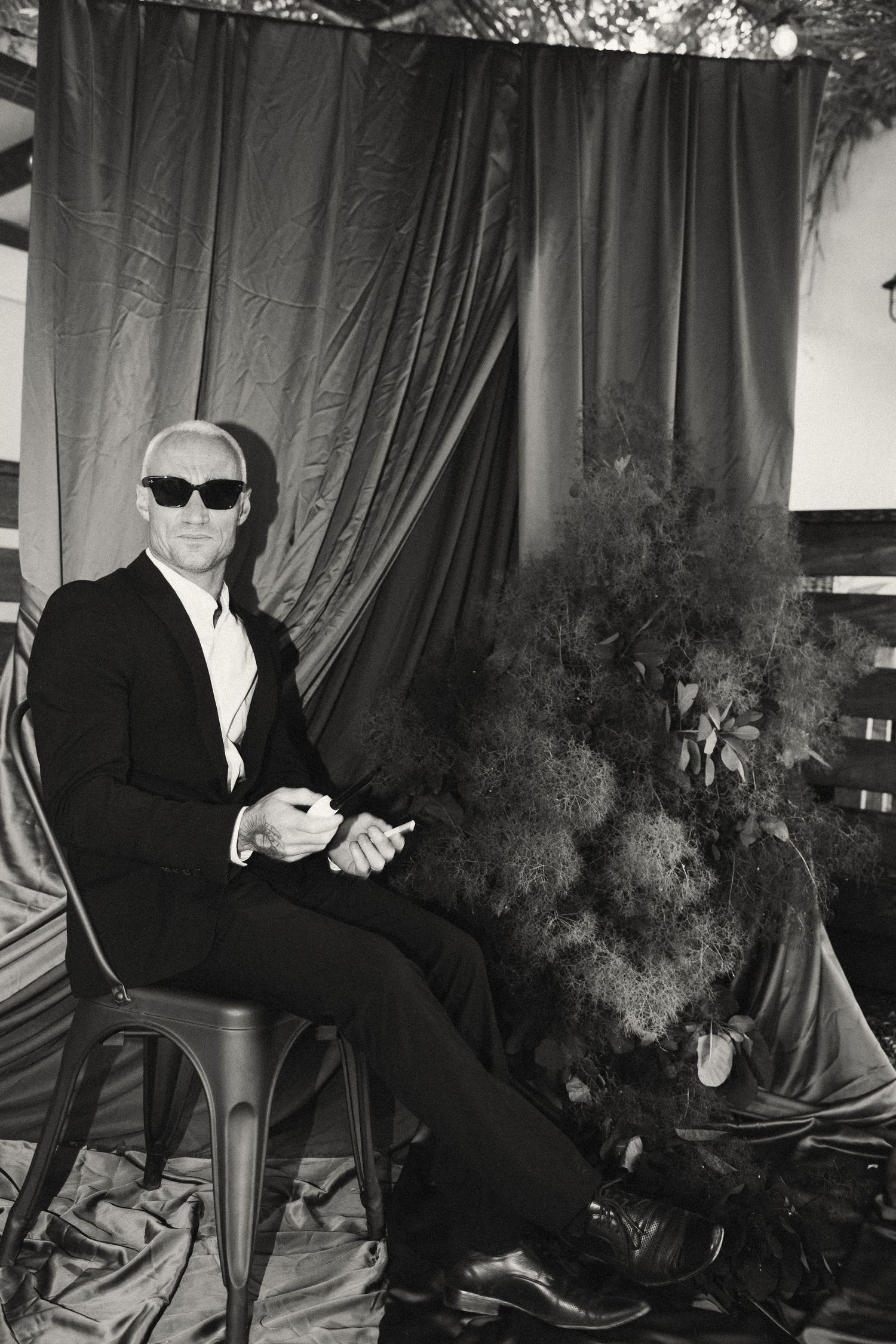 Figure in sunglasses and suit seated against dark curtain backdrop.