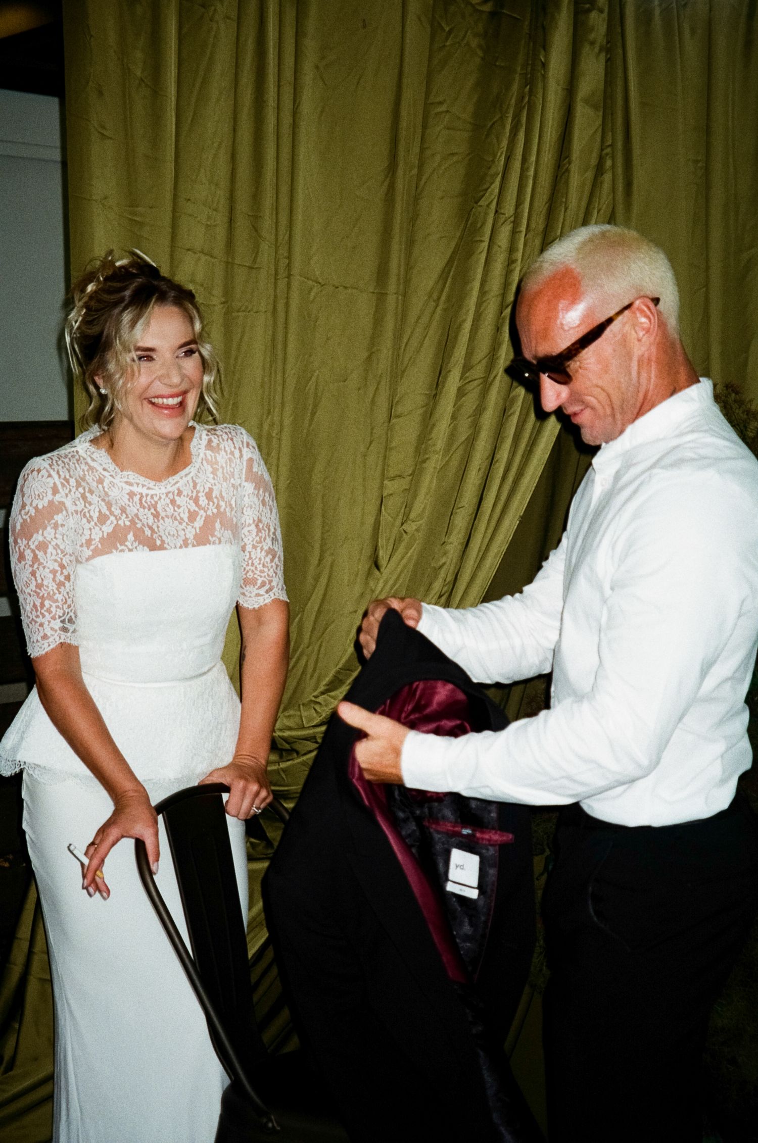 A bride in a white lace wedding dress and an older man in a white suit interact backstage at an event.