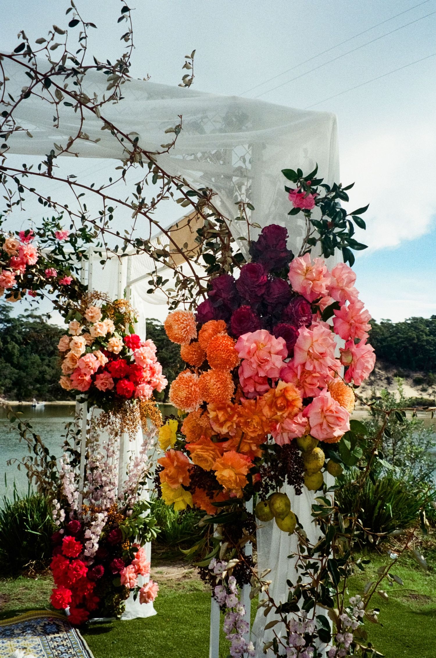 Vibrant floral arch arrangement featuring orange, pink, and burgundy blooms against a lakeside outdoor setting.