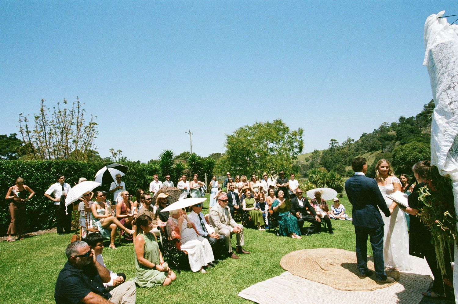 Panoramic view of an outdoor wedding ceremony on a grassy hilltop with guests seated in white chairs.