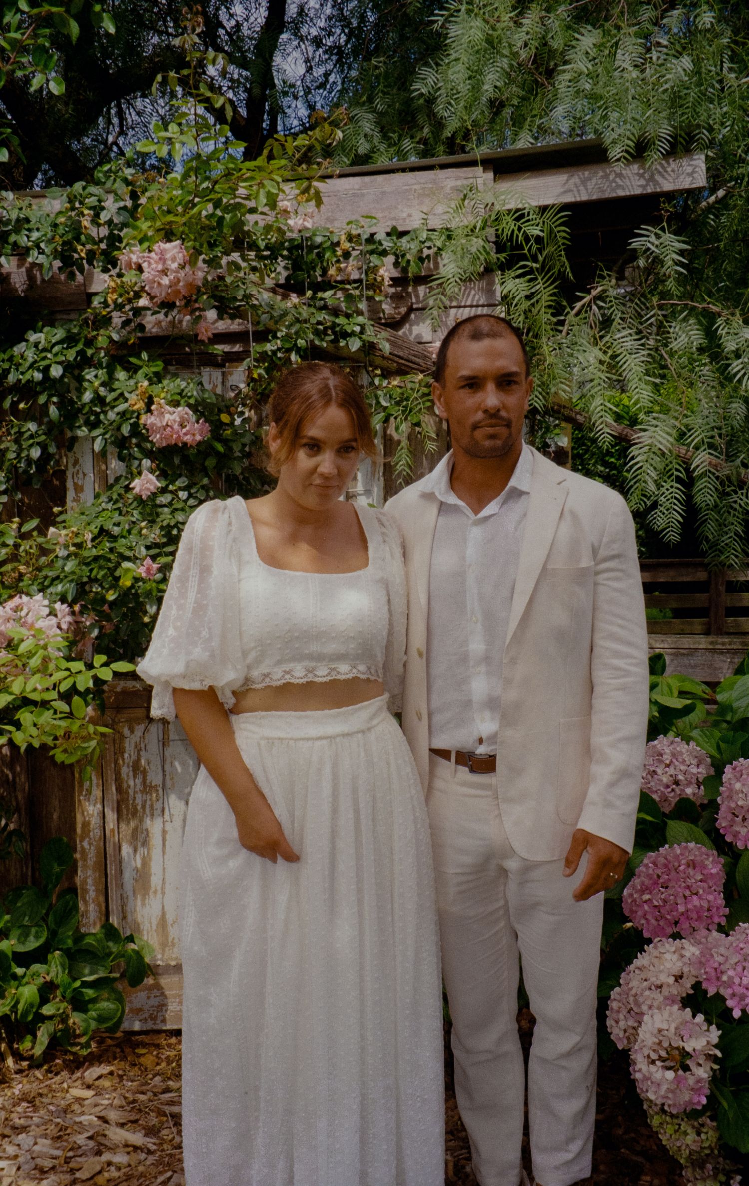 A couple poses in white outfits in a lush garden setting with pink flowers and greenery.
