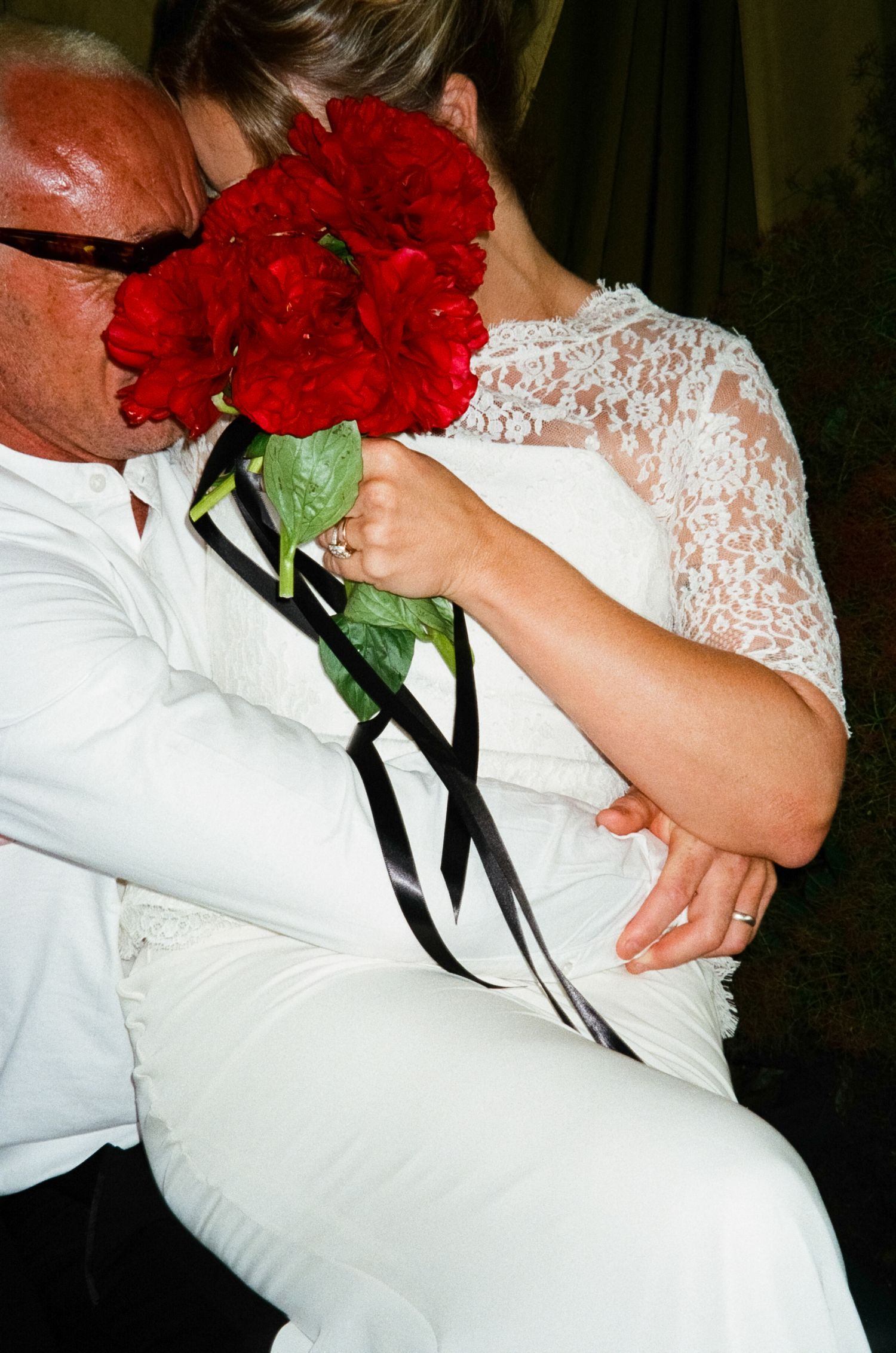 A romantic moment captured with red roses and white lace as a couple embraces at what appears to be a wedding celebration.