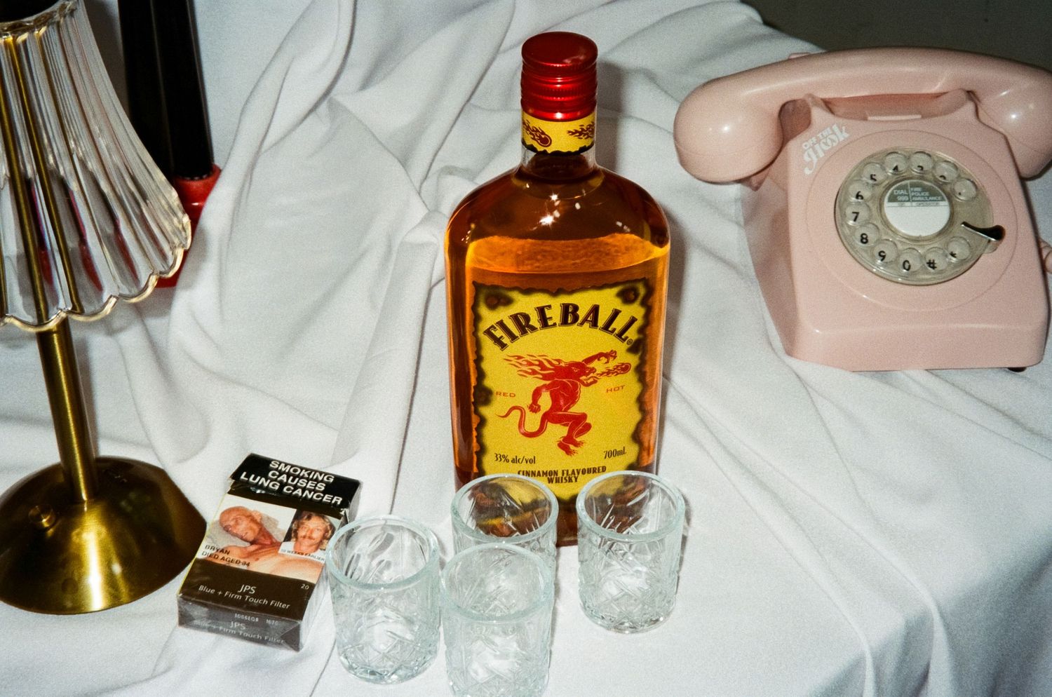 A bottle of Fireball whiskey sits next to a vintage pink rotary telephone and some crystal glasses on a white surface.