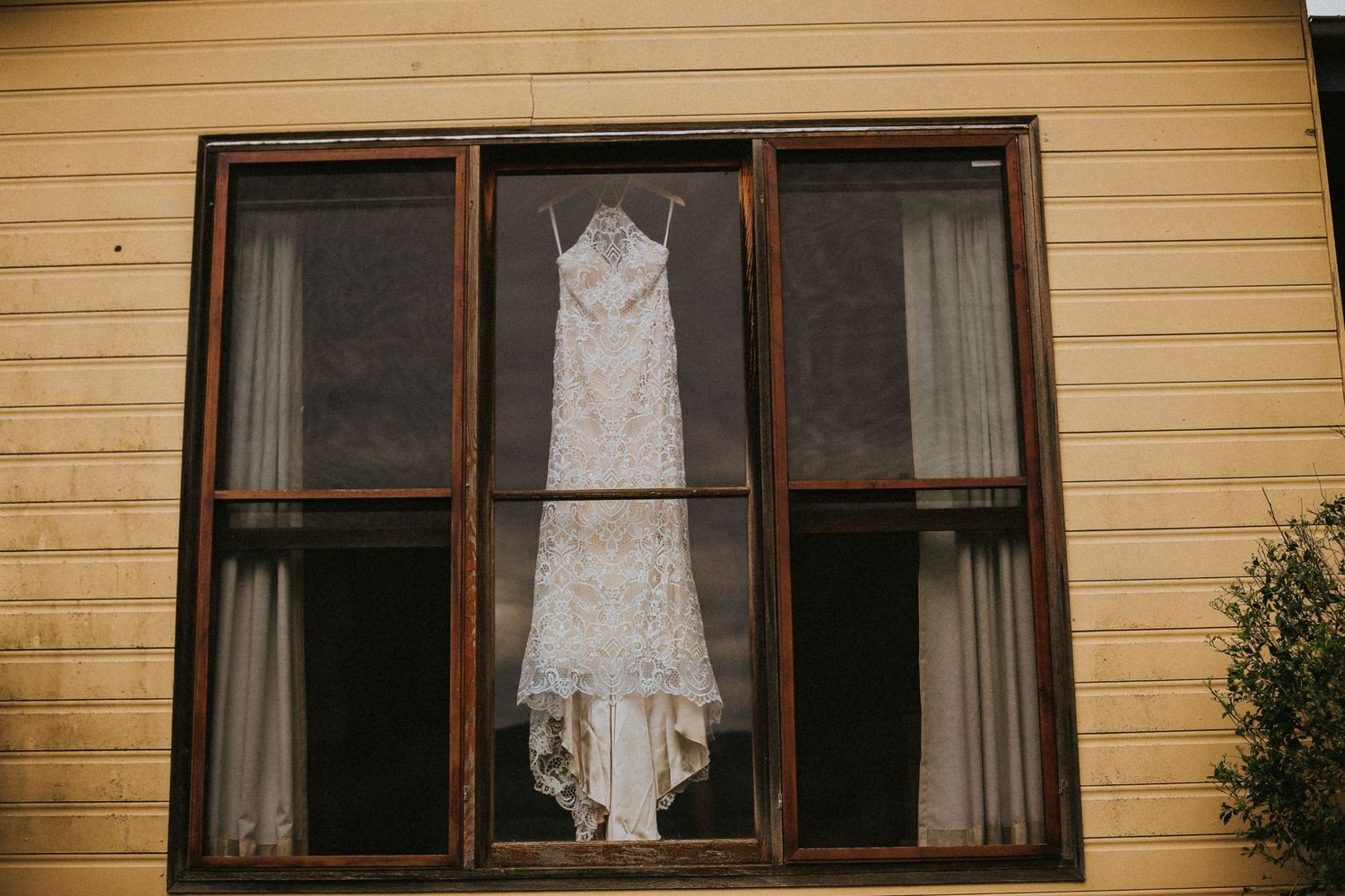 A white lace wedding dress hangs in a window of a yellow house, displayed between dark curtains.