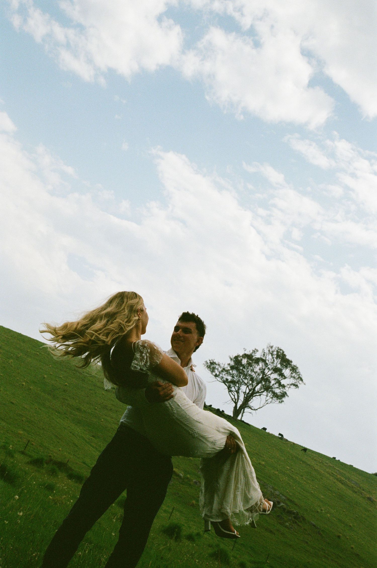 A couple spins joyfully together on a grassy hillside under a bright blue sky.