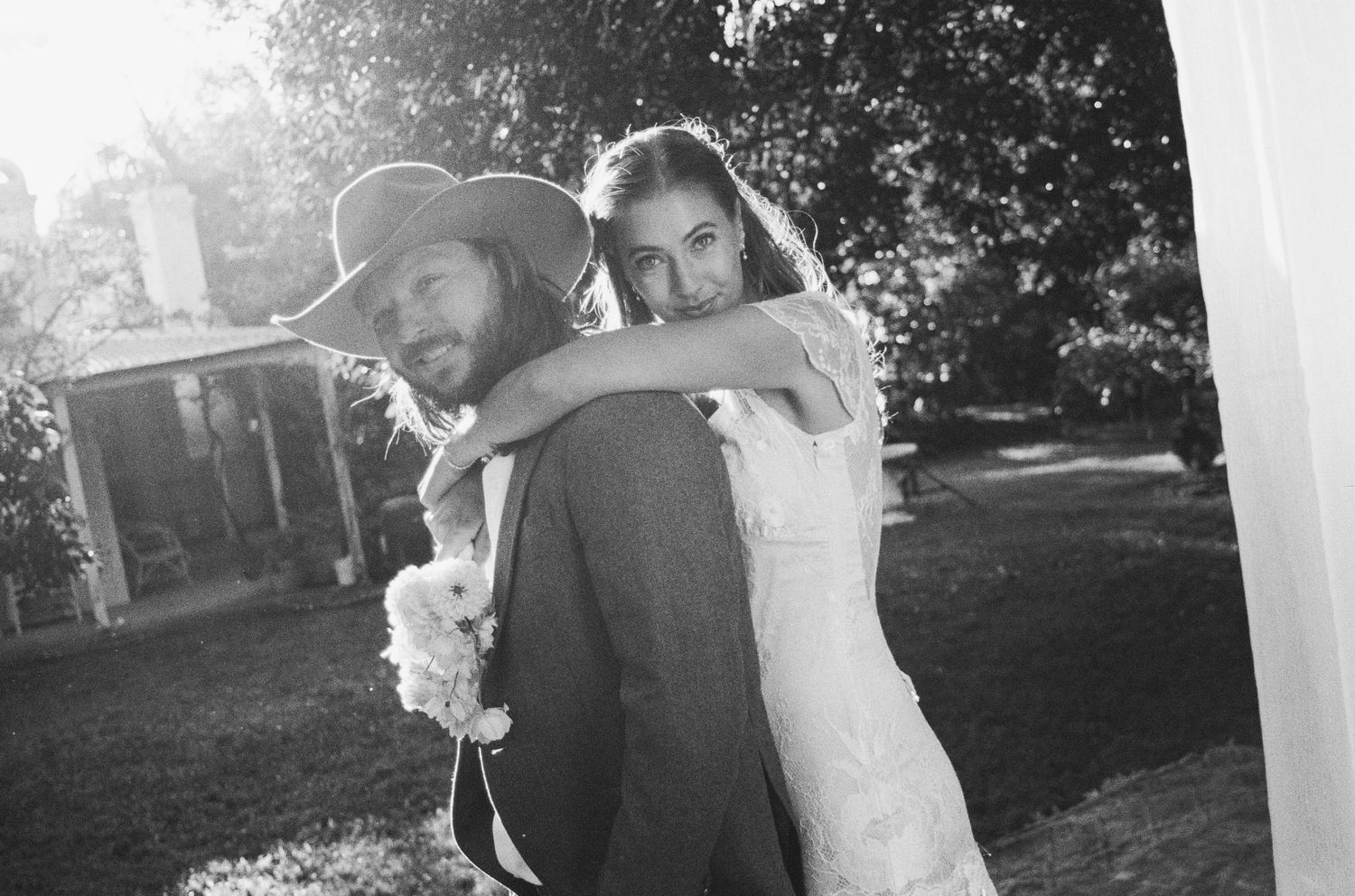 Black and white photo of a bride hugging a groom wearing a cowboy hat outdoors at a wedding.