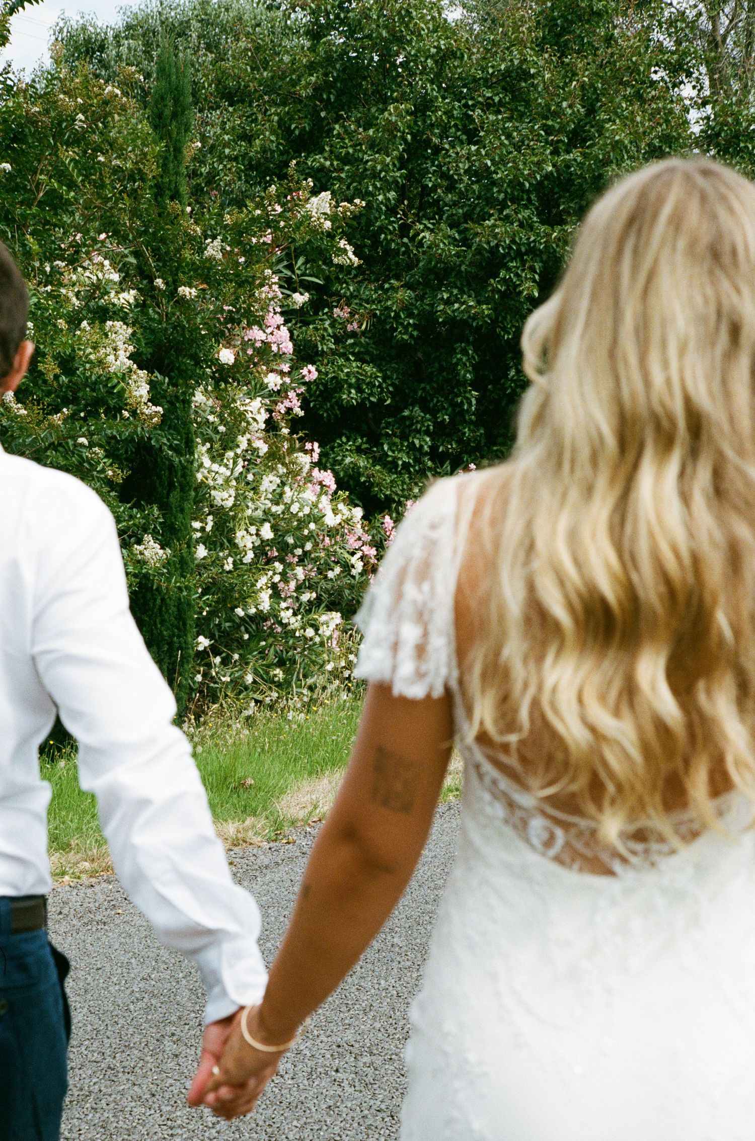 A couple holding hands on a path surrounded by blooming flowers and greenery.