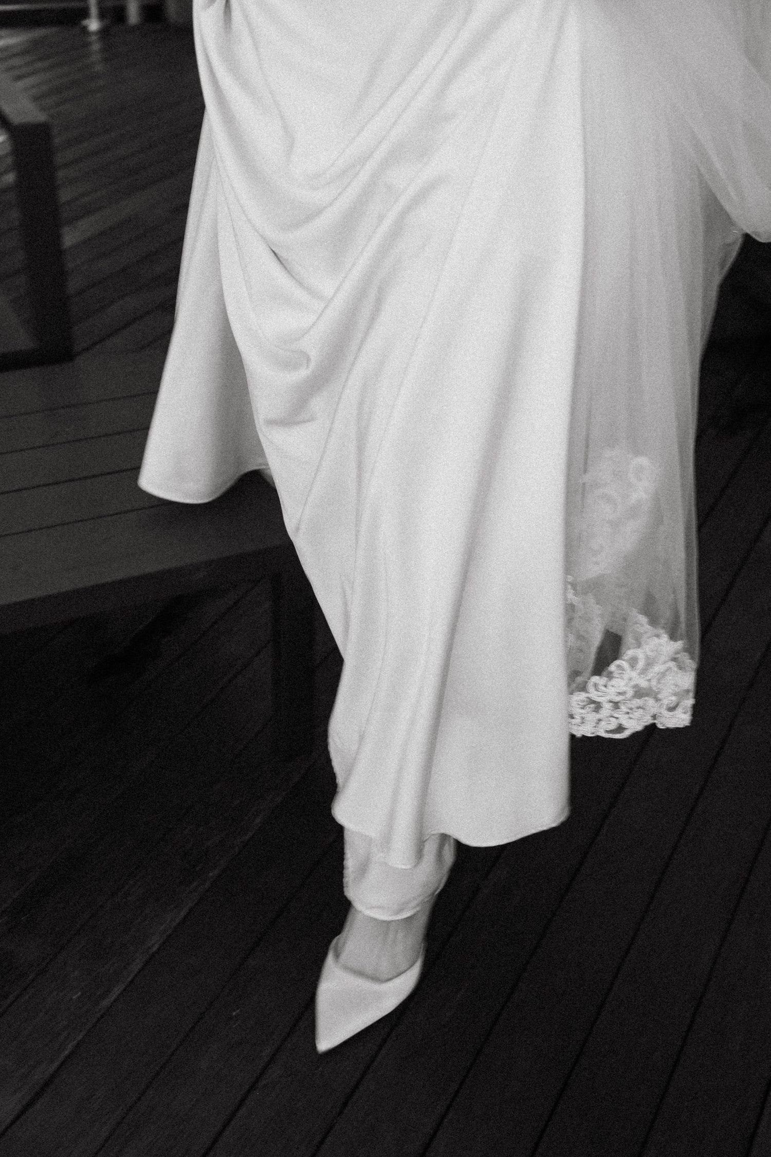 Close-up details of a flowing white wedding dress and shoes on a dark wooden floor.