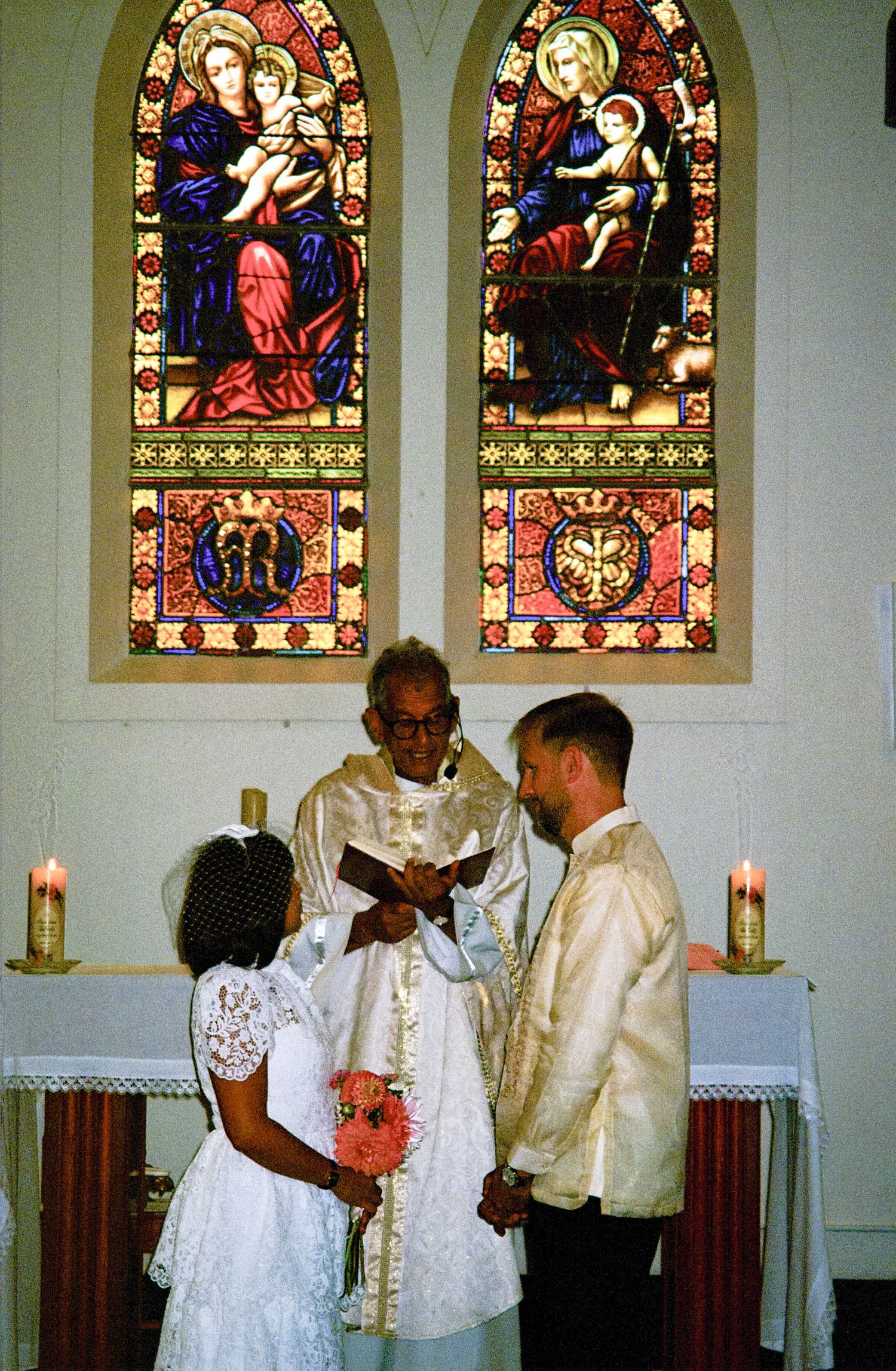 Traditional wedding ceremony taking place in front of stained glass church windows depicting religious iconography.