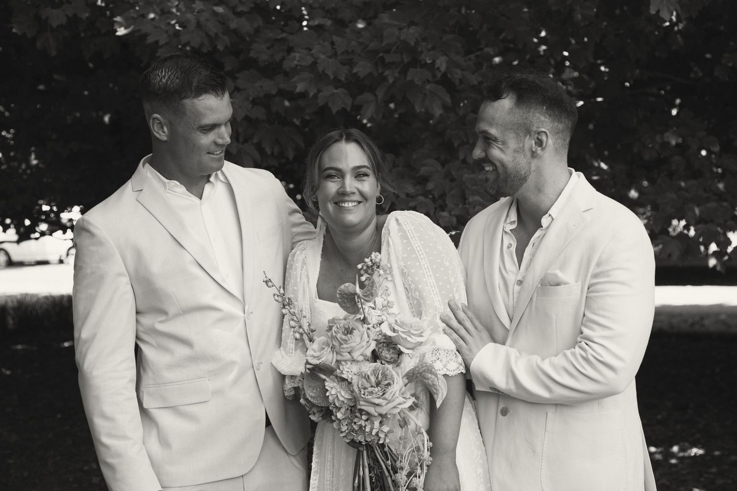 A black and white wedding photo showing three people outdoors with the bride holding a bouquet and wearing white.
