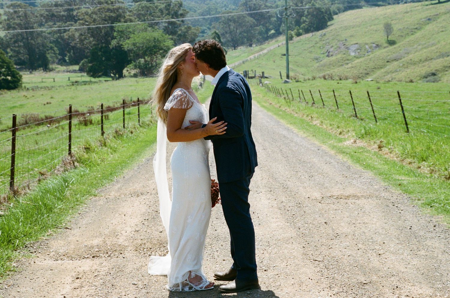 A couple shares a romantic moment on a dirt road surrounded by rolling green hills and pastures.