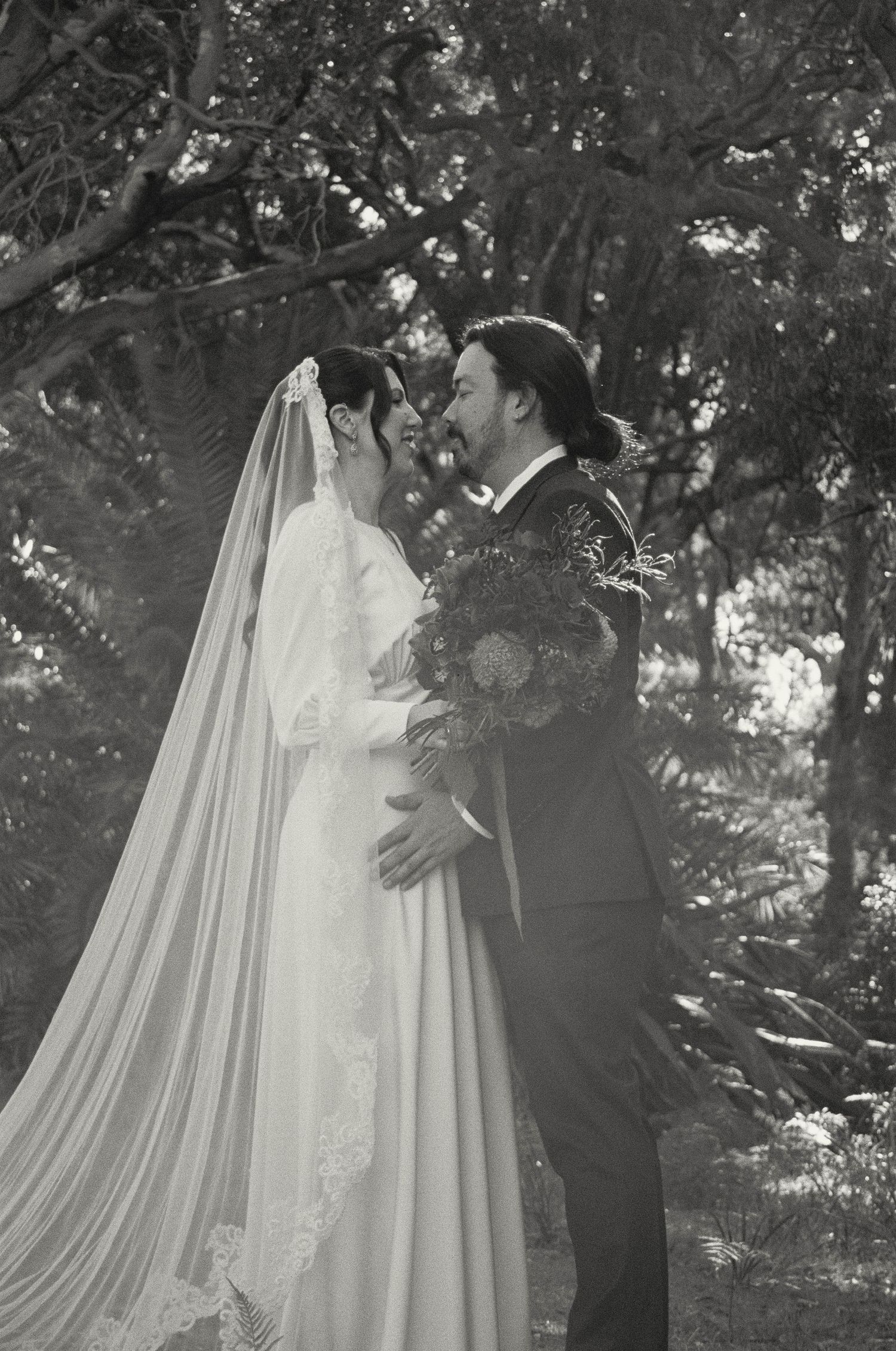 A romantic black and white wedding photo of a couple sharing an intimate moment under trees in an outdoor setting.