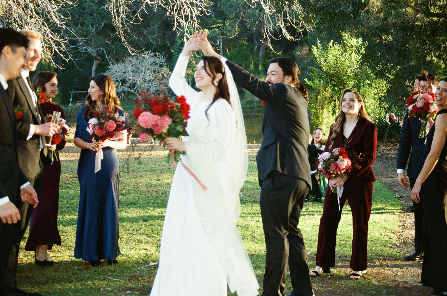 Wedding party arranged in formation on grassy lawn with evergreen trees in background during summer celebration.