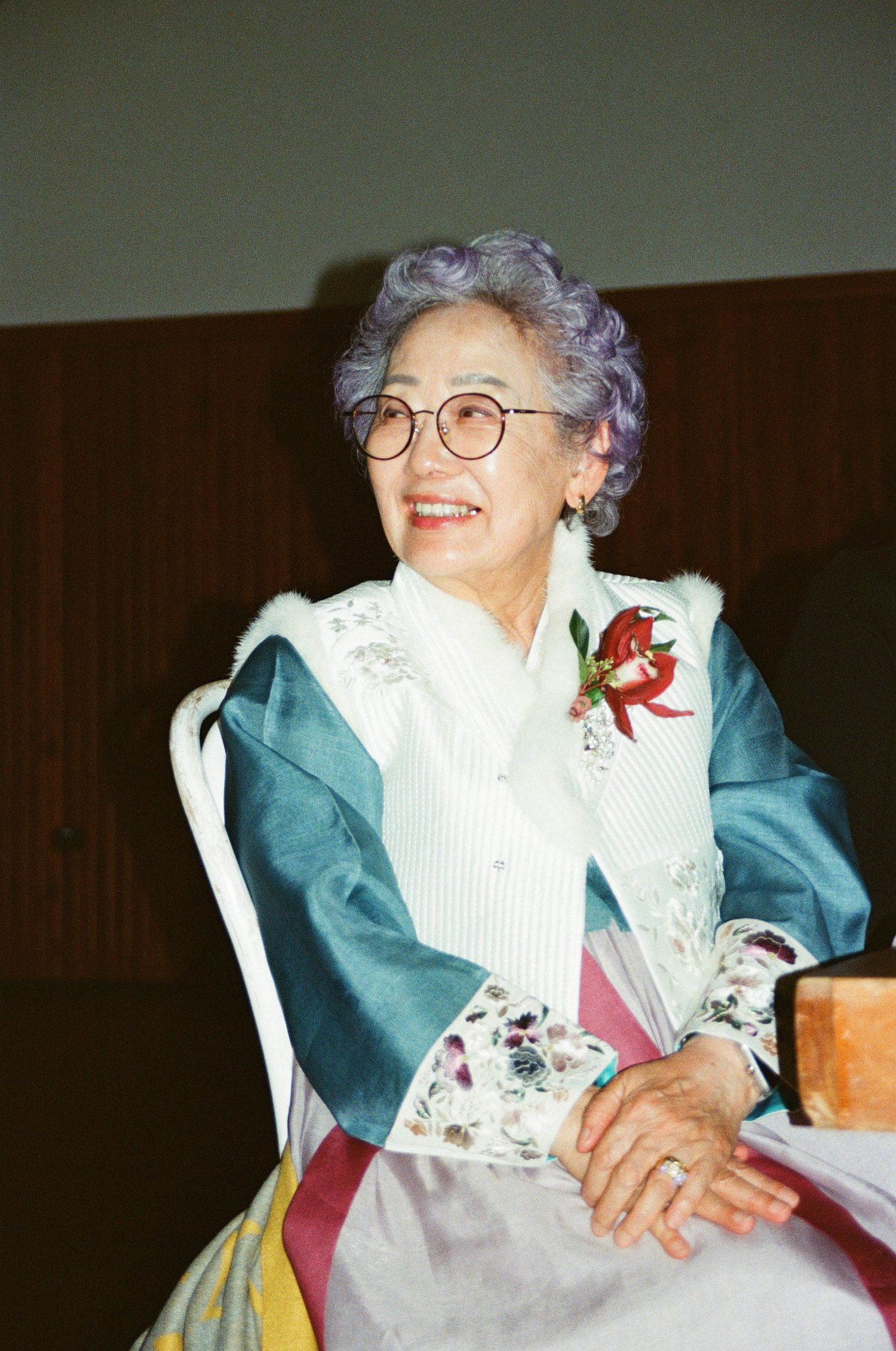 An elderly woman wearing traditional Korean hanbok and glasses sits smiling with a corsage pin.