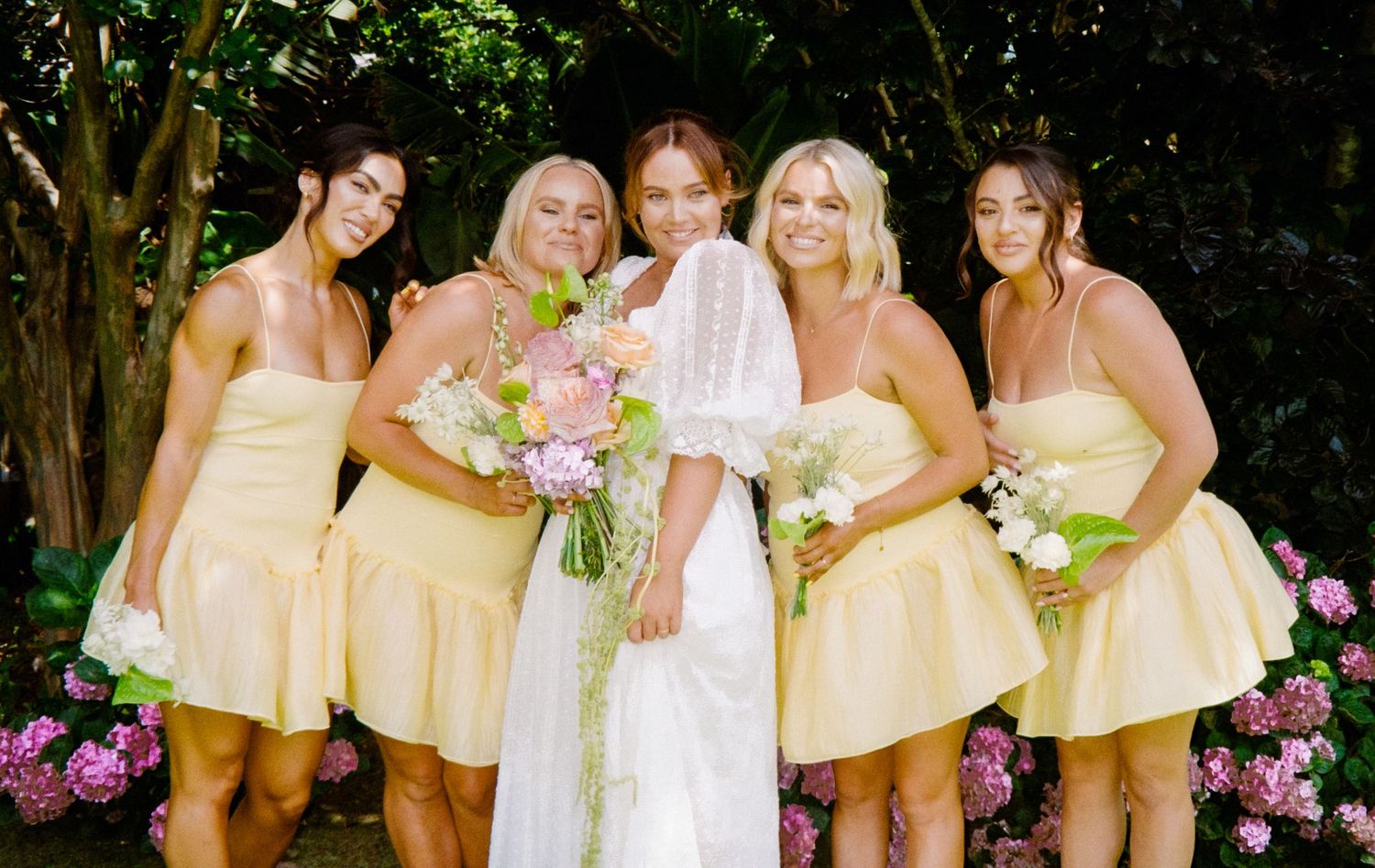 A bridal party poses outdoors in pastel yellow bridesmaid dresses with pink floral bouquets against a garden backdrop.