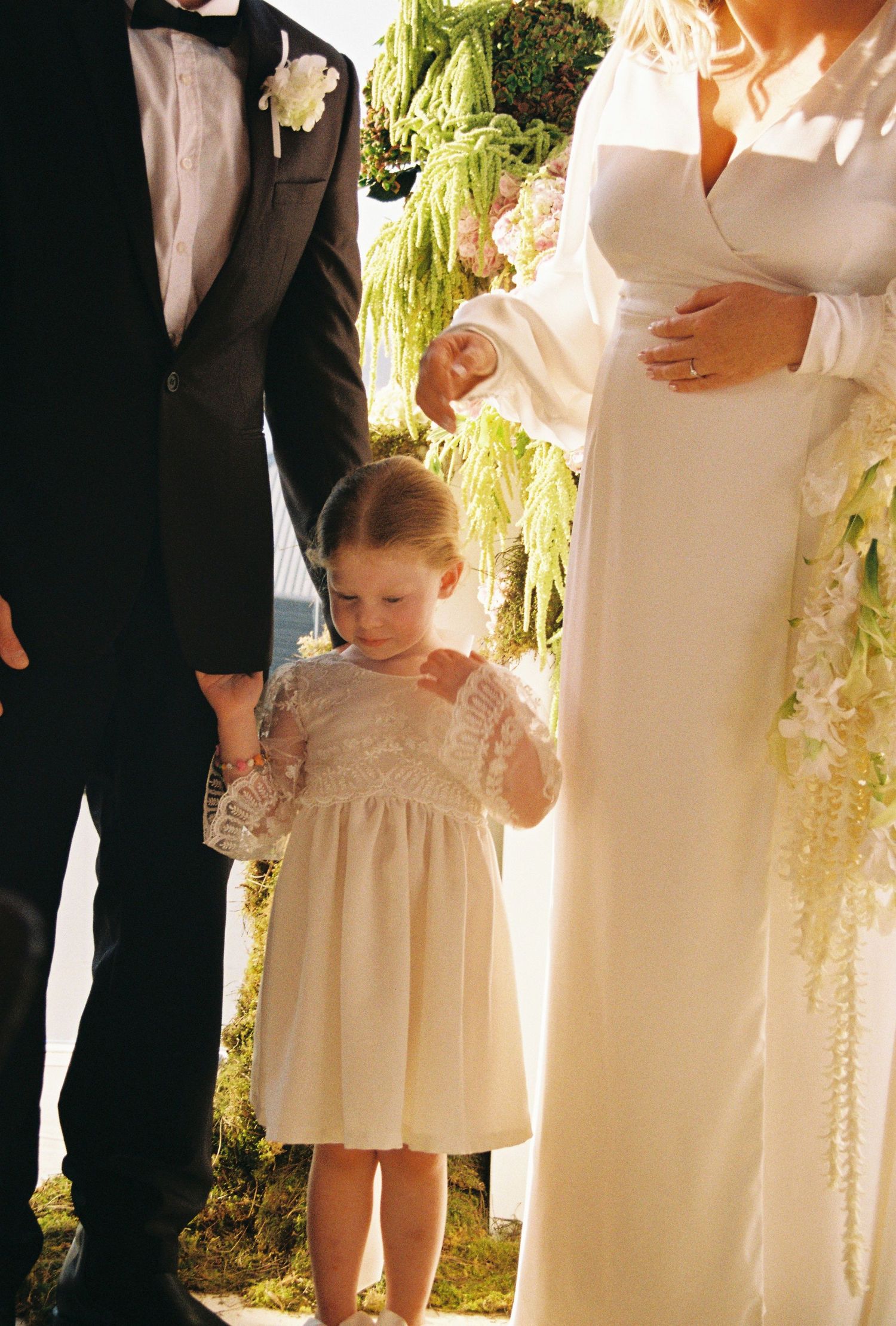 A small child in a white dress stands next to formal wedding attire in an outdoor garden setting.