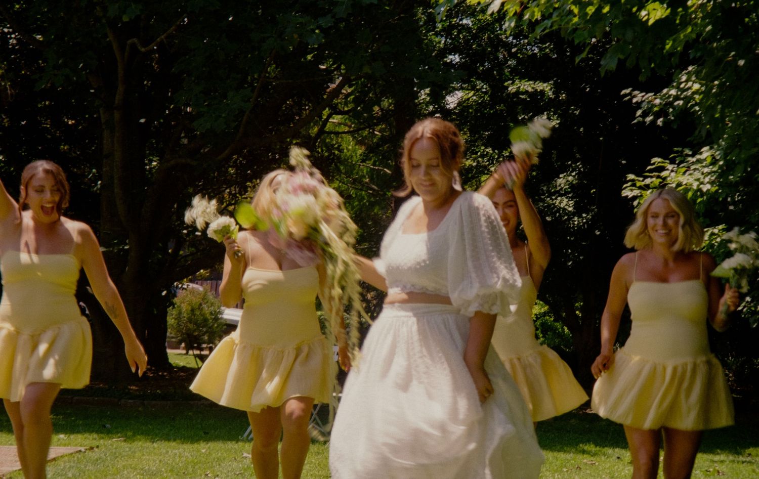 A wedding party in pastel yellow bridesmaid dresses walks together through a sunny garden setting.