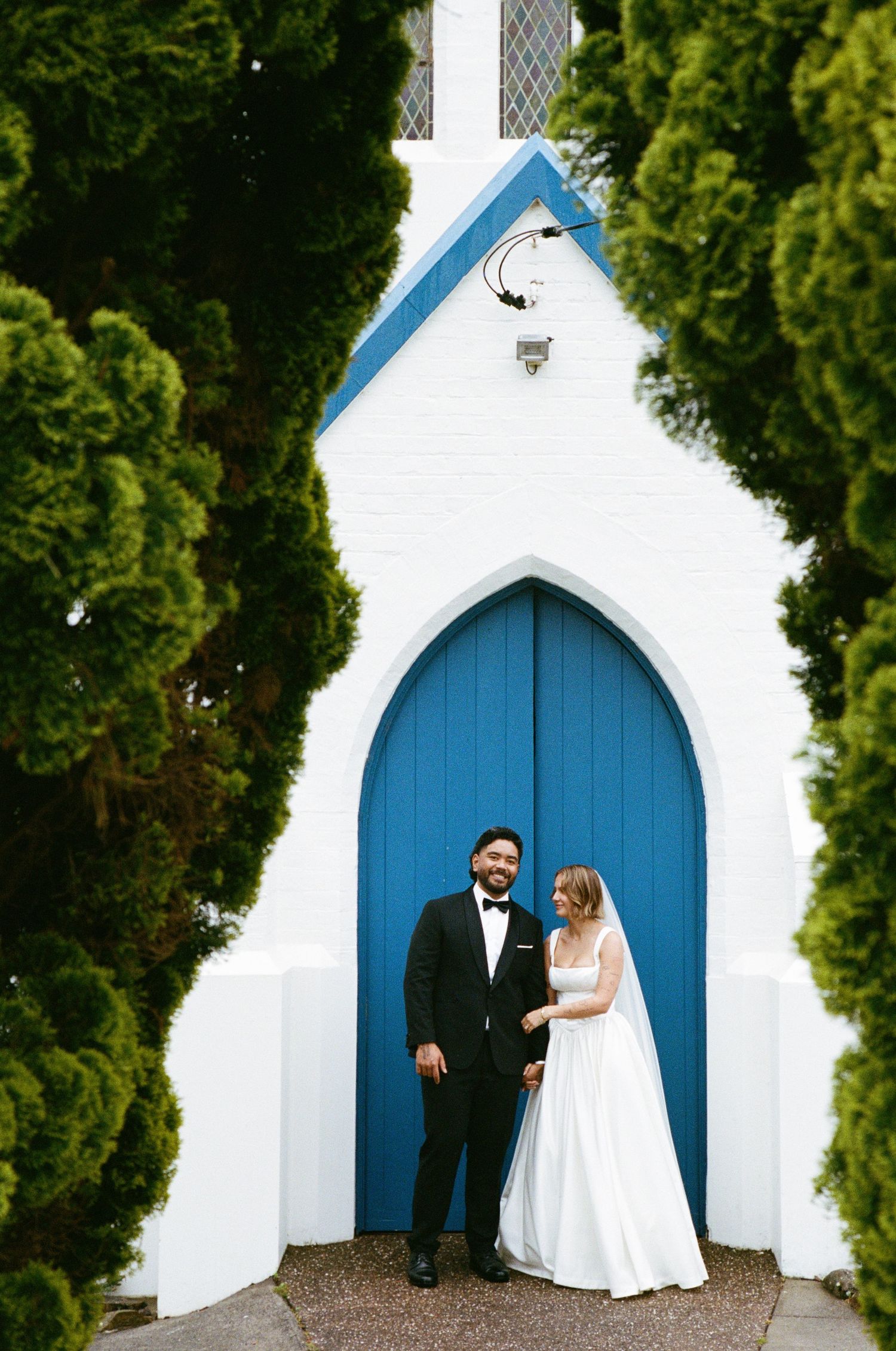 A blue church door serves as a backdrop for a wedding photo session in front of white walls and green foliage.