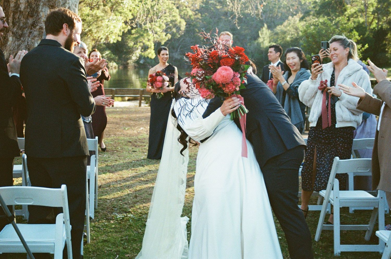 Outdoor garden wedding ceremony with abundant red flowers and white folding chairs.