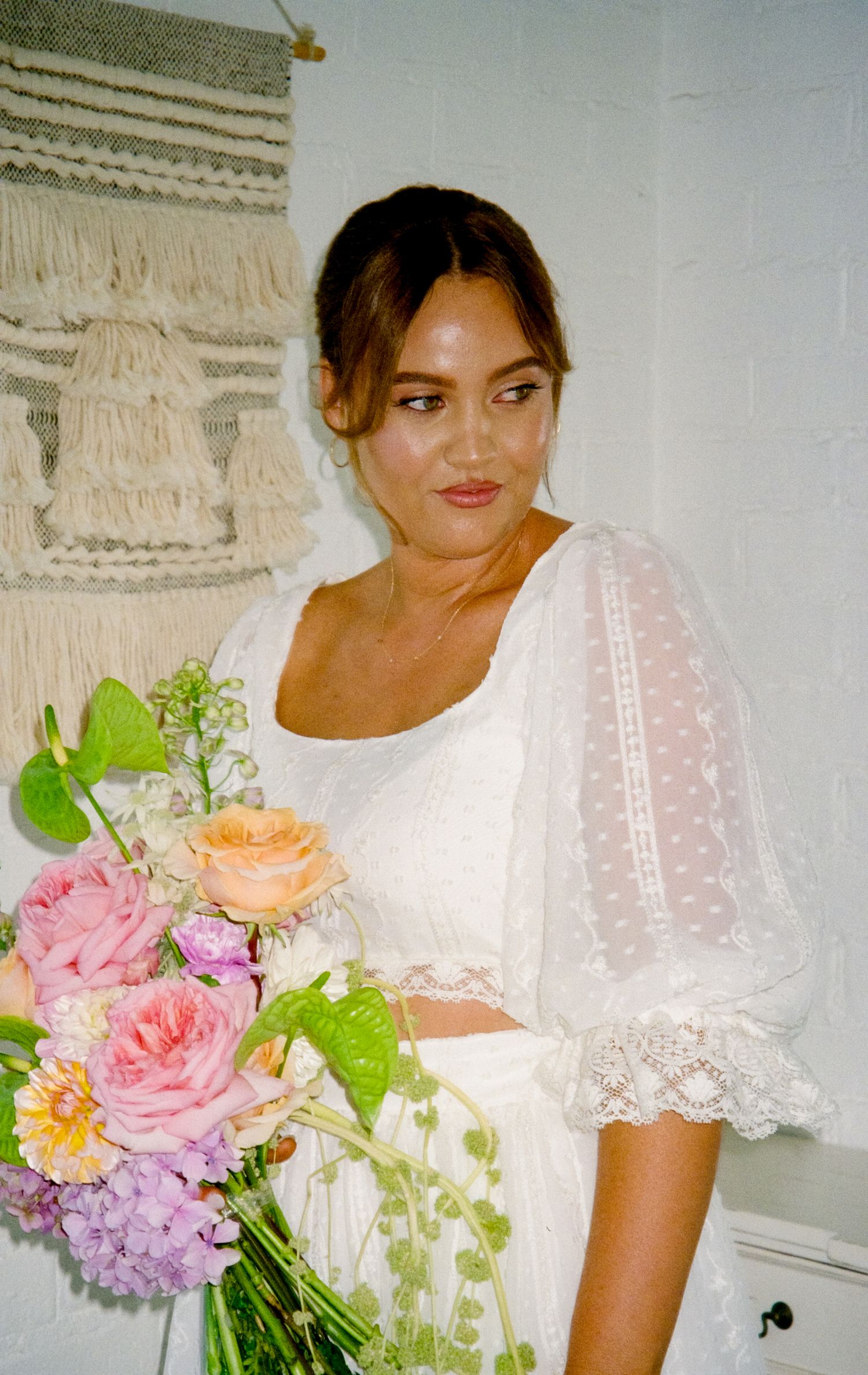 Person in white lace dress holding a colorful bouquet of pink, peach and purple flowers against white wall.