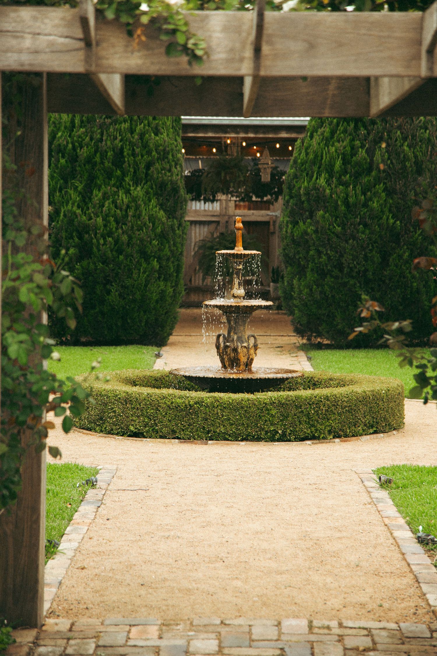 Elegant garden path leads to ornate tiered fountain surrounded by manicured hedges and string lights overhead.