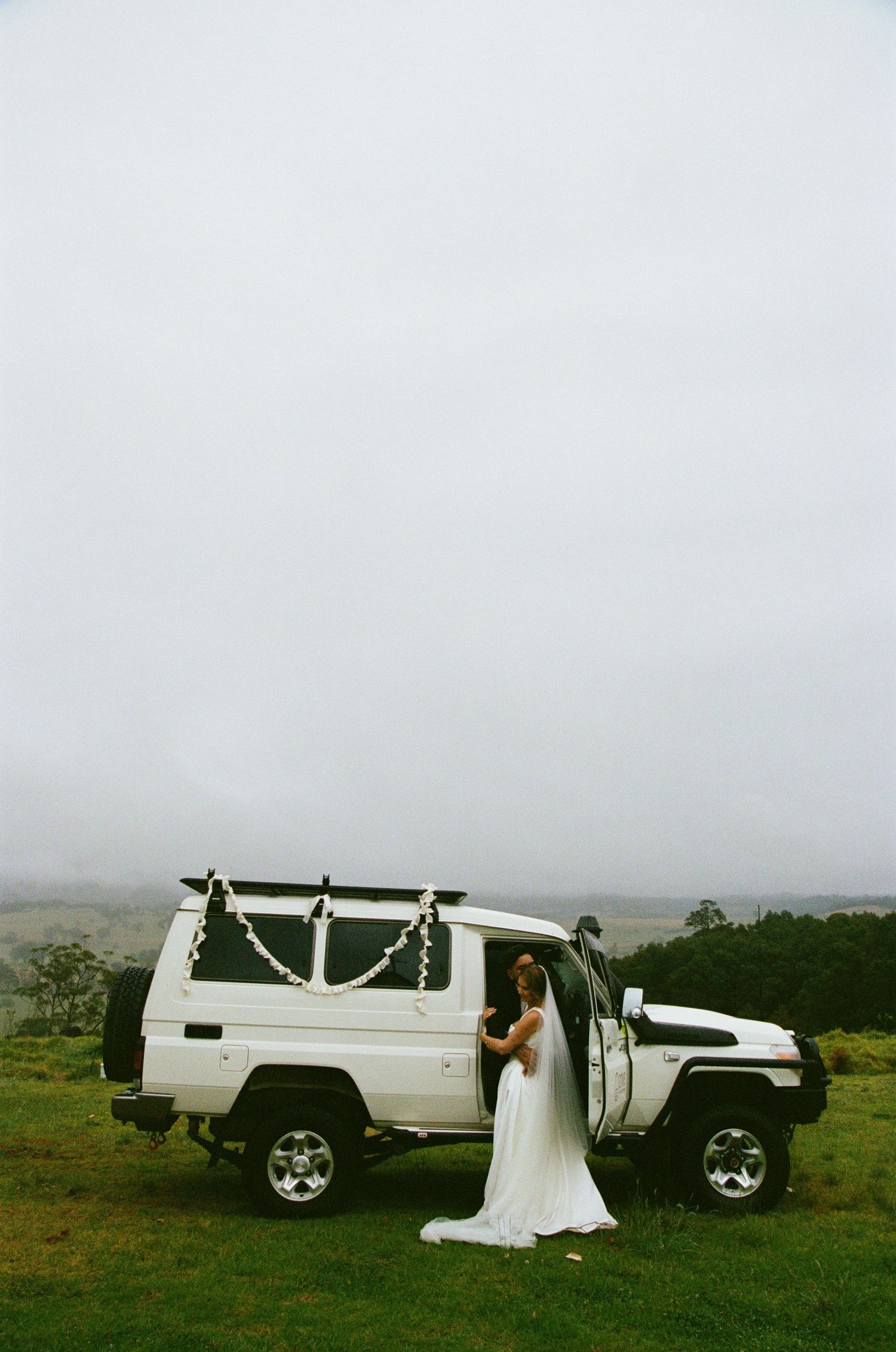 A bride in white stands beside a decorated white Land Rover on misty terrain.