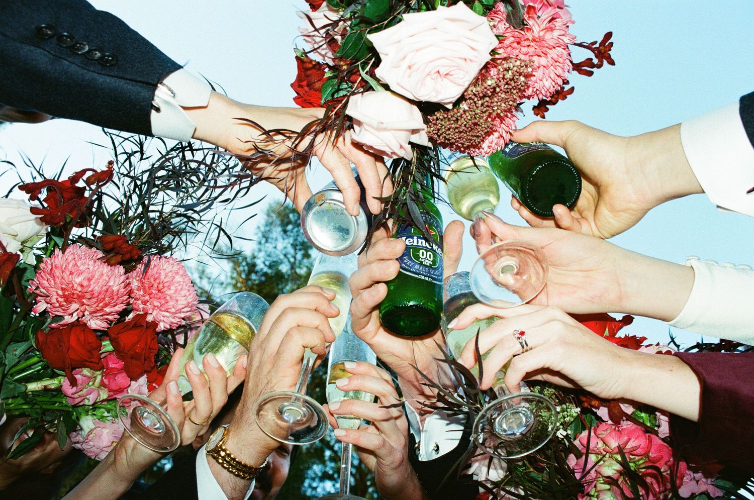 Multiple hands raise glasses for a celebratory toast surrounded by pink and white floral arrangements.