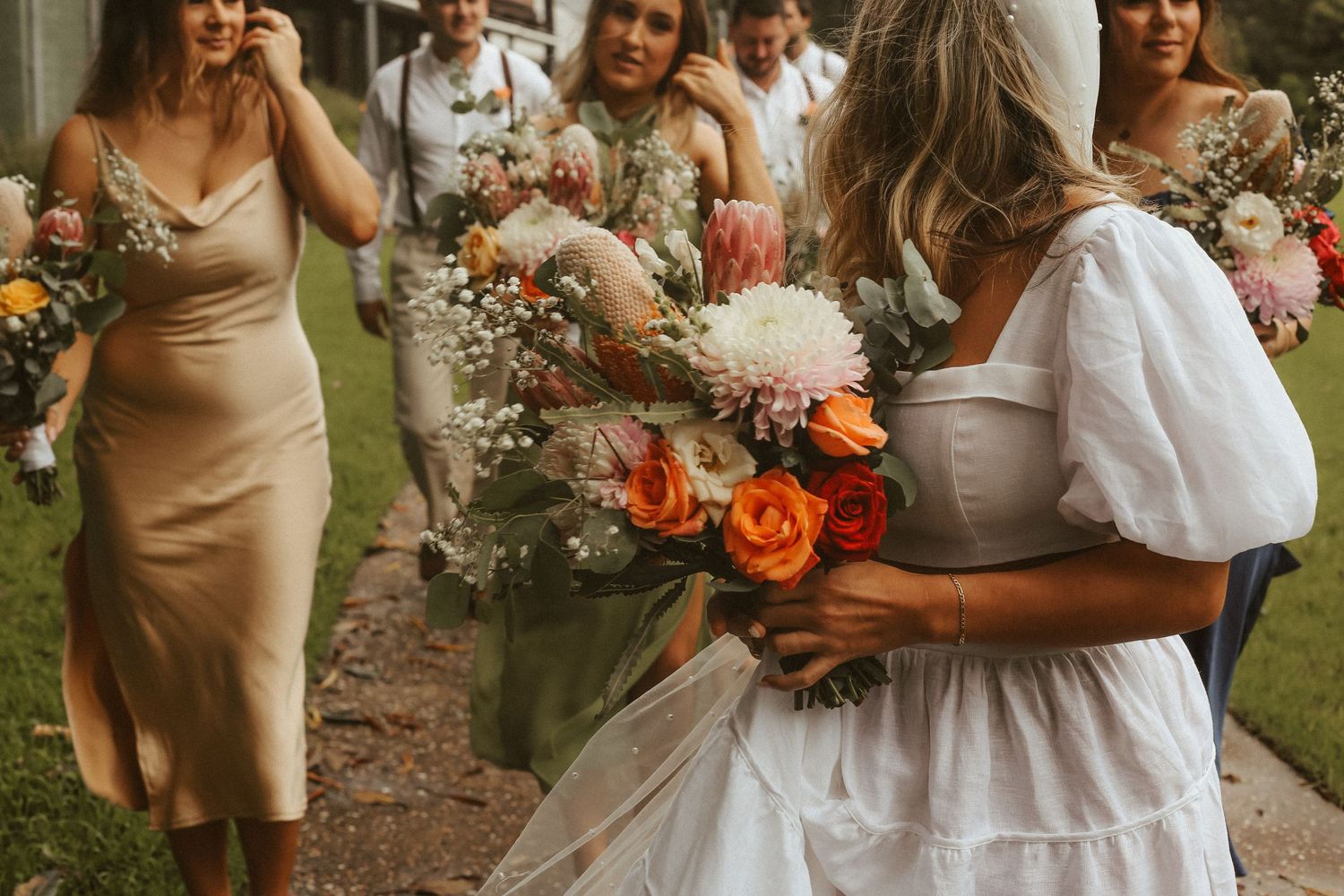 Group of bridesmaids holding vibrant orange and pink bouquets pose in champagne-colored dresses at outdoor wedding.