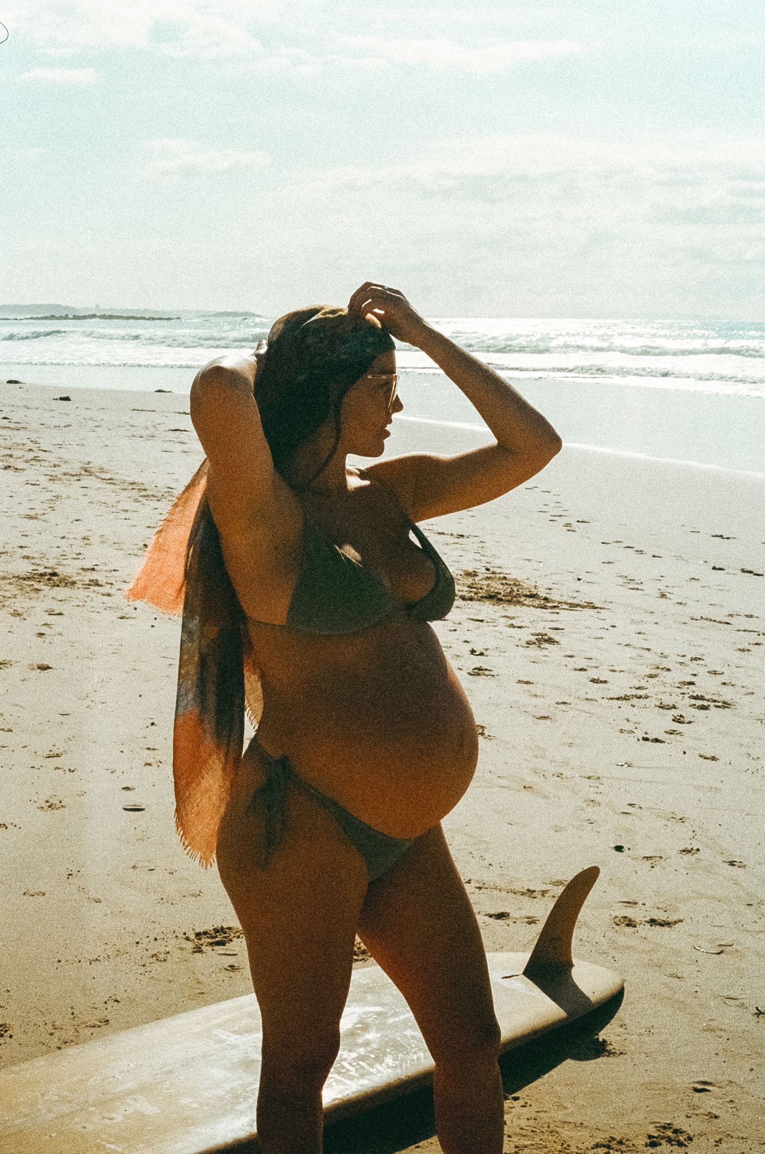 Pregnant woman in bikini holding surfboard on sunny beach, wearing headscarf, ocean waves in background.