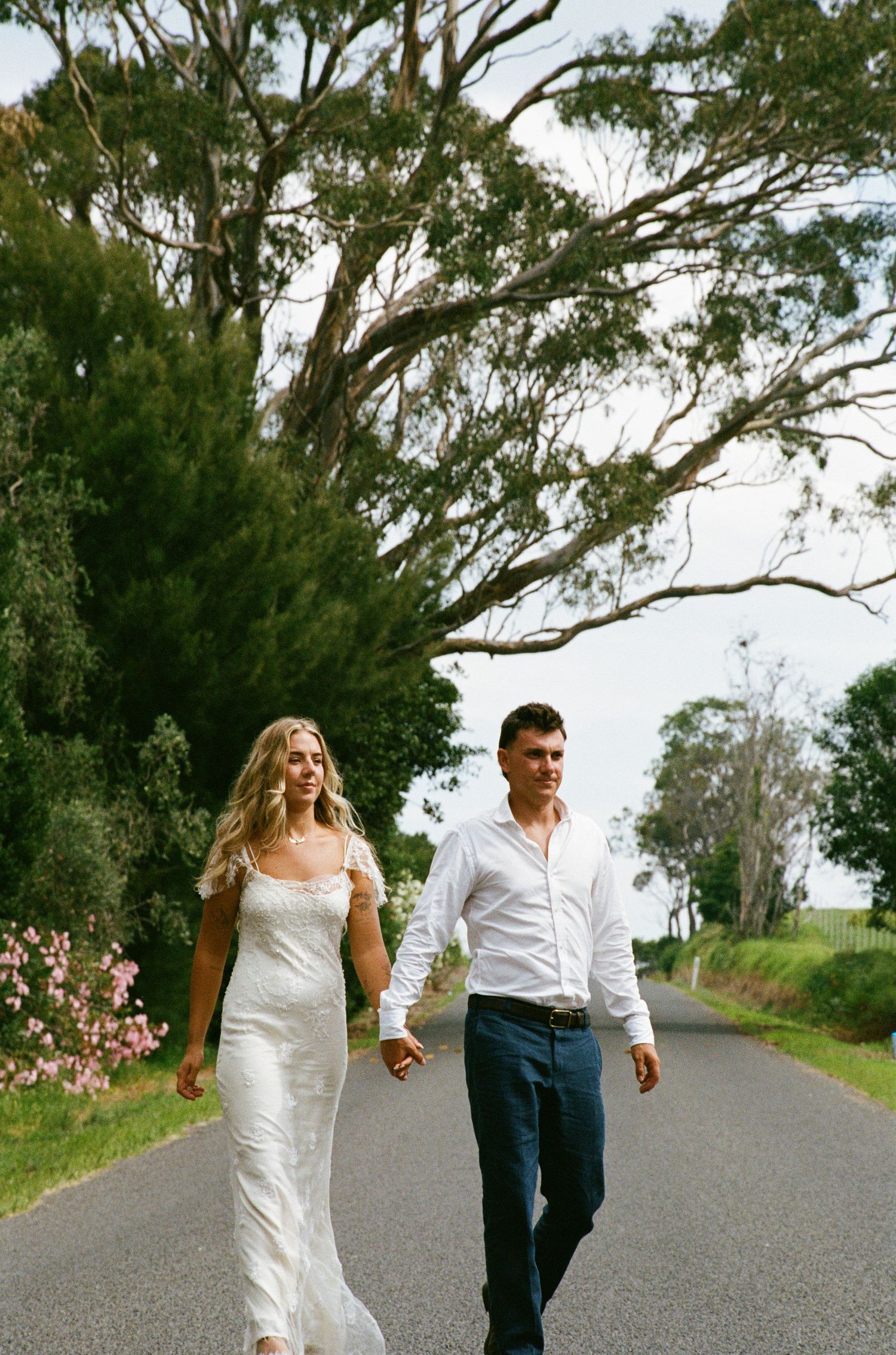 A couple walks hand in hand down a tree-lined country road, with the bride wearing a flowing white dress.