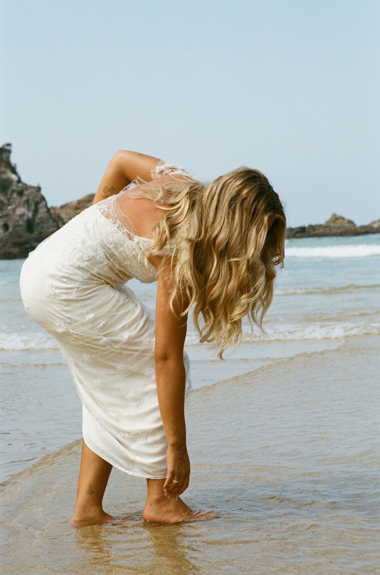 Person in a white dress bending down to touch the ocean waves at the beach on a sunny day.