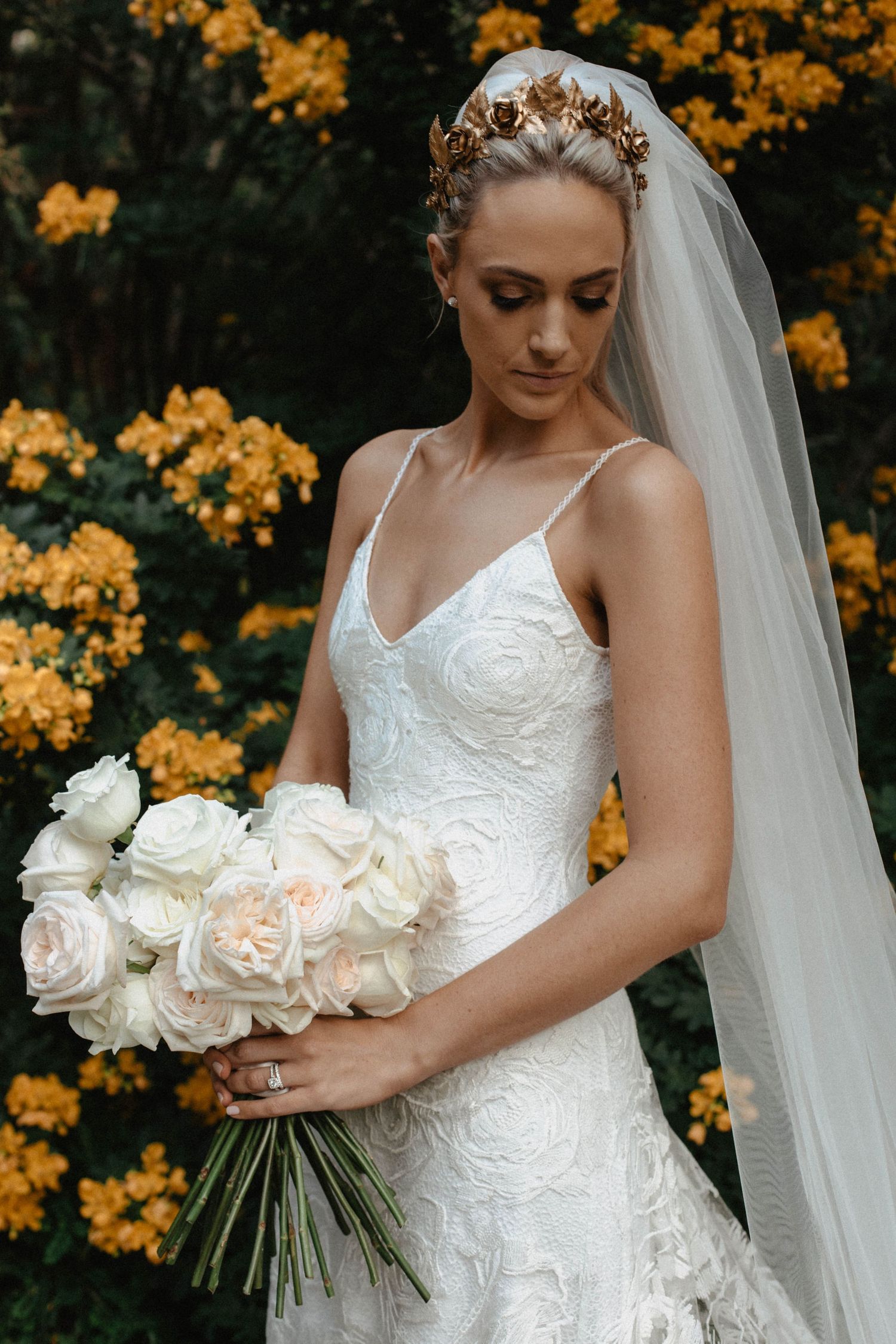 Bride in lace wedding dress holds white rose bouquet against backdrop of golden autumn foliage.