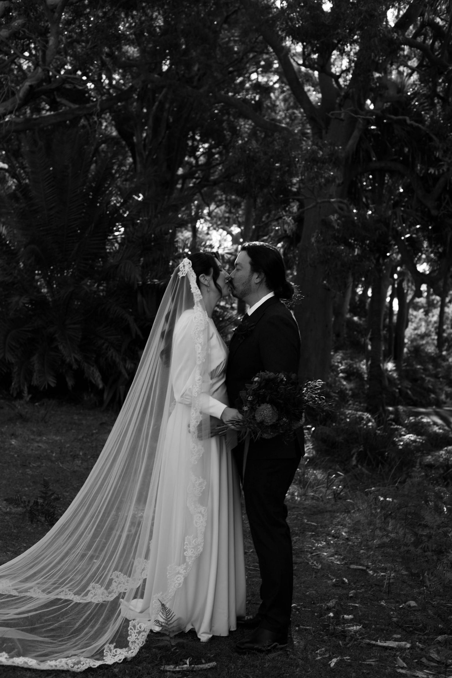 Black and white photo of couple in wedding attire embracing under trees with long flowing veil.