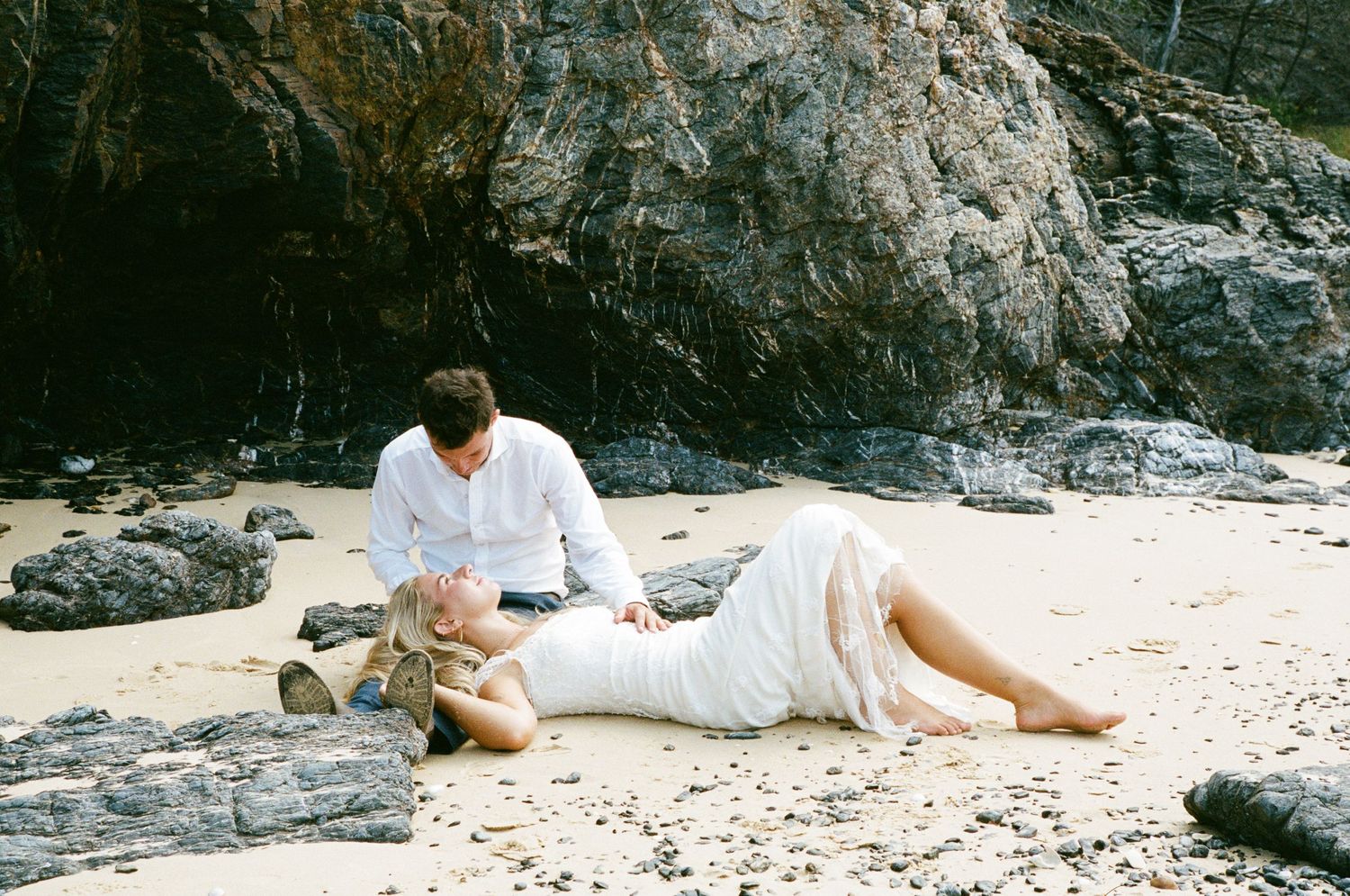 Someone in white clothing relaxes while reading a book on a rocky beach by the ocean.
