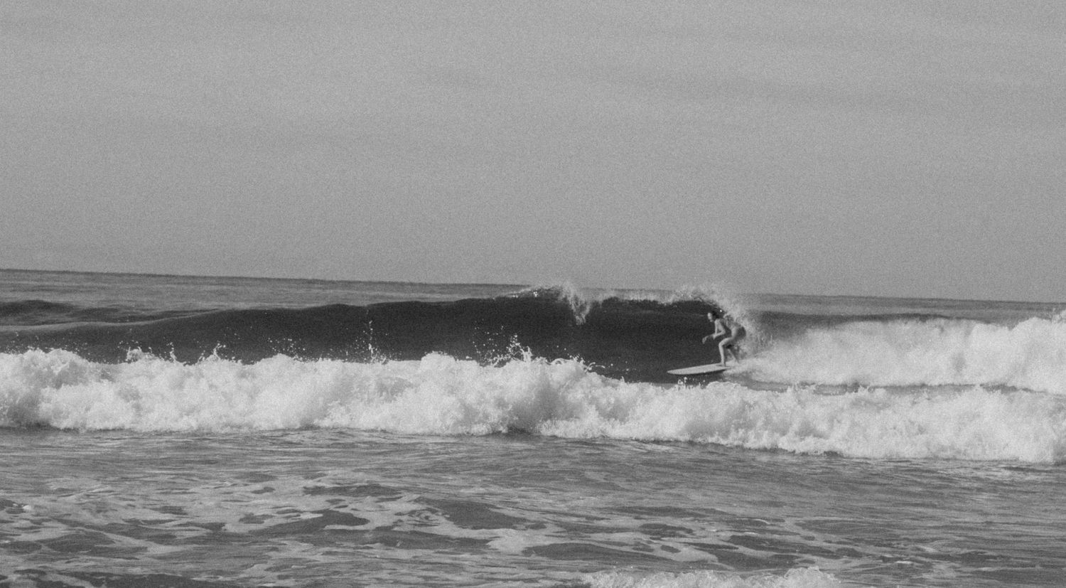 A surfer rides a dark wave in black and white as ocean spray crashes behind them.