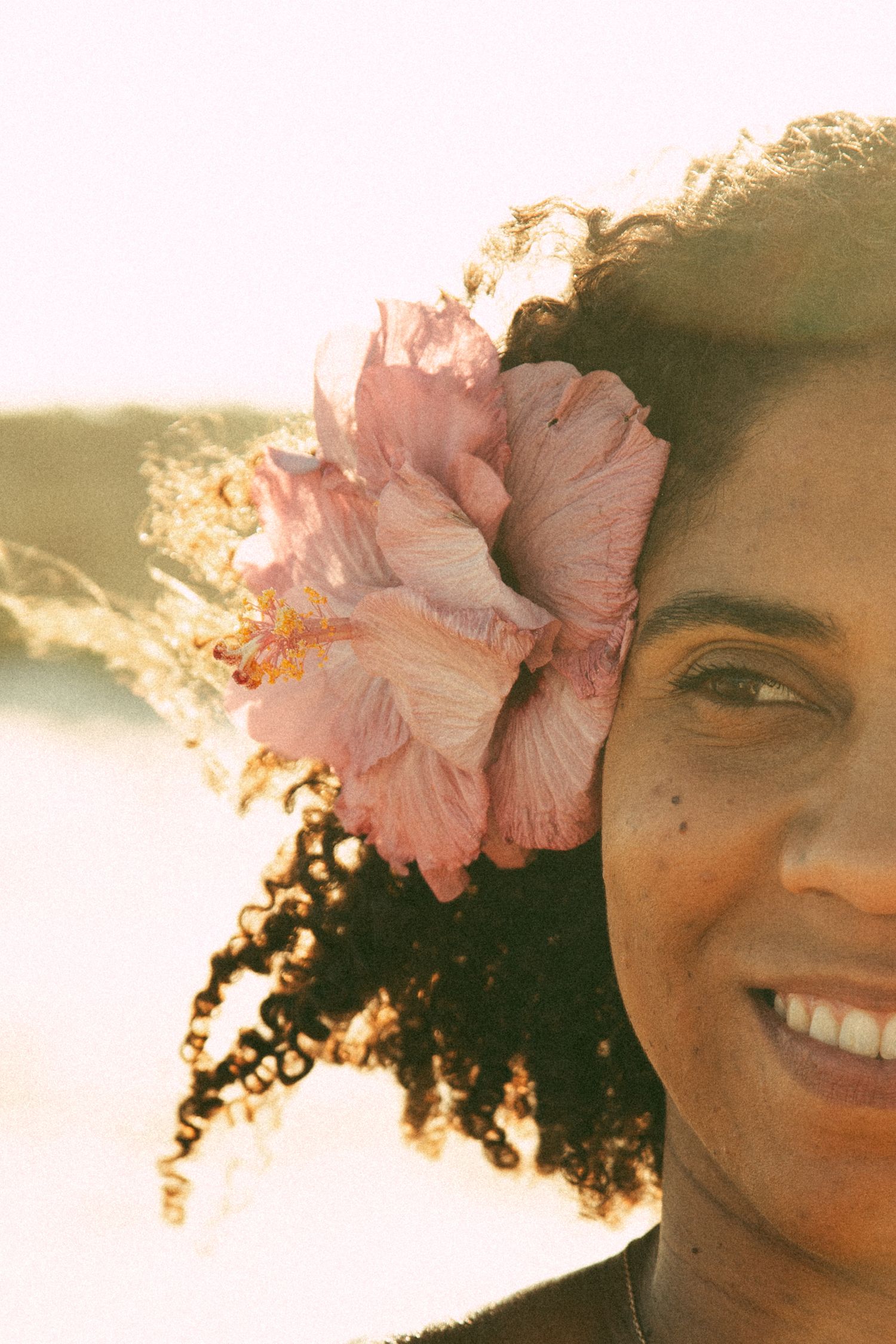 Woman with curly hair wearing pink hibiscus flower in golden sunset light.