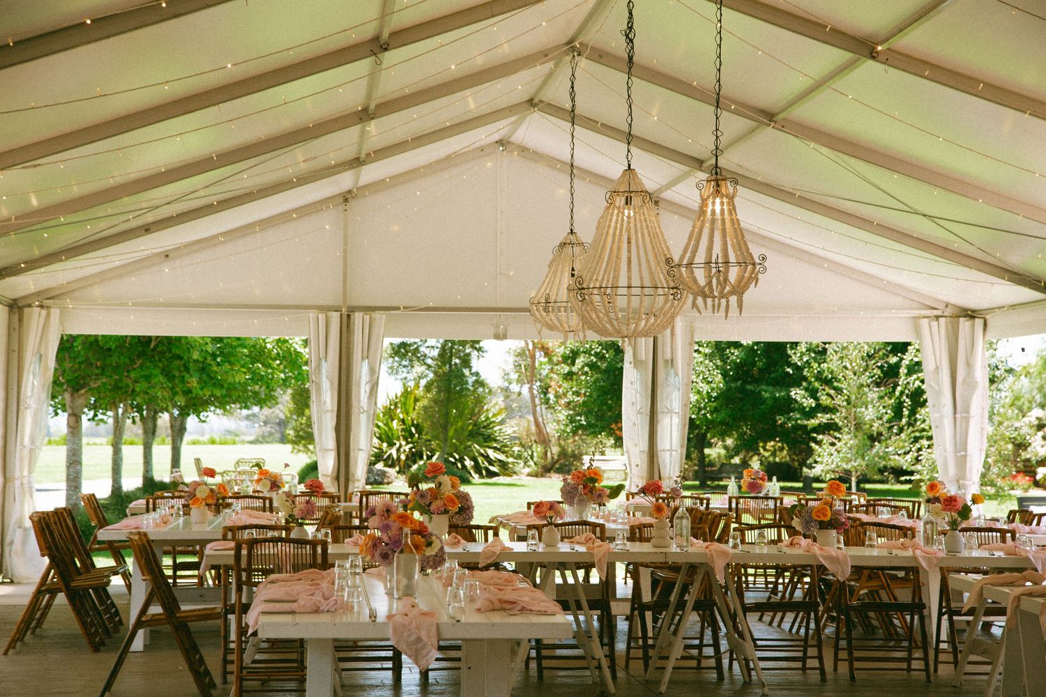 Wedding reception tent setup with rustic wooden tables and hanging chandeliers in an outdoor setting.