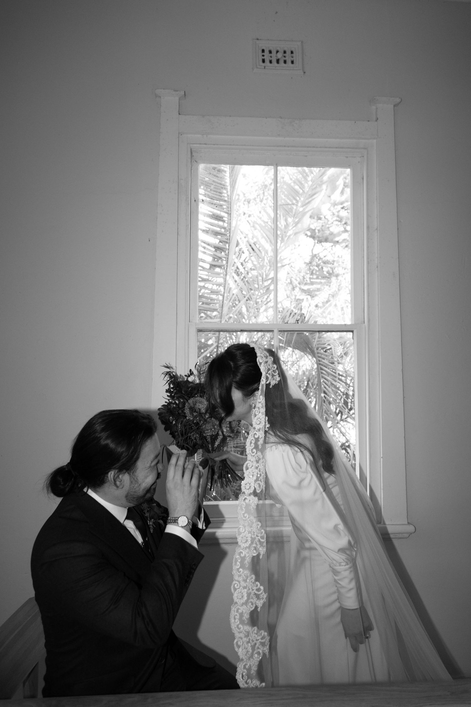 Wedding couple shares intimate moment by vintage window in black and white photograph.