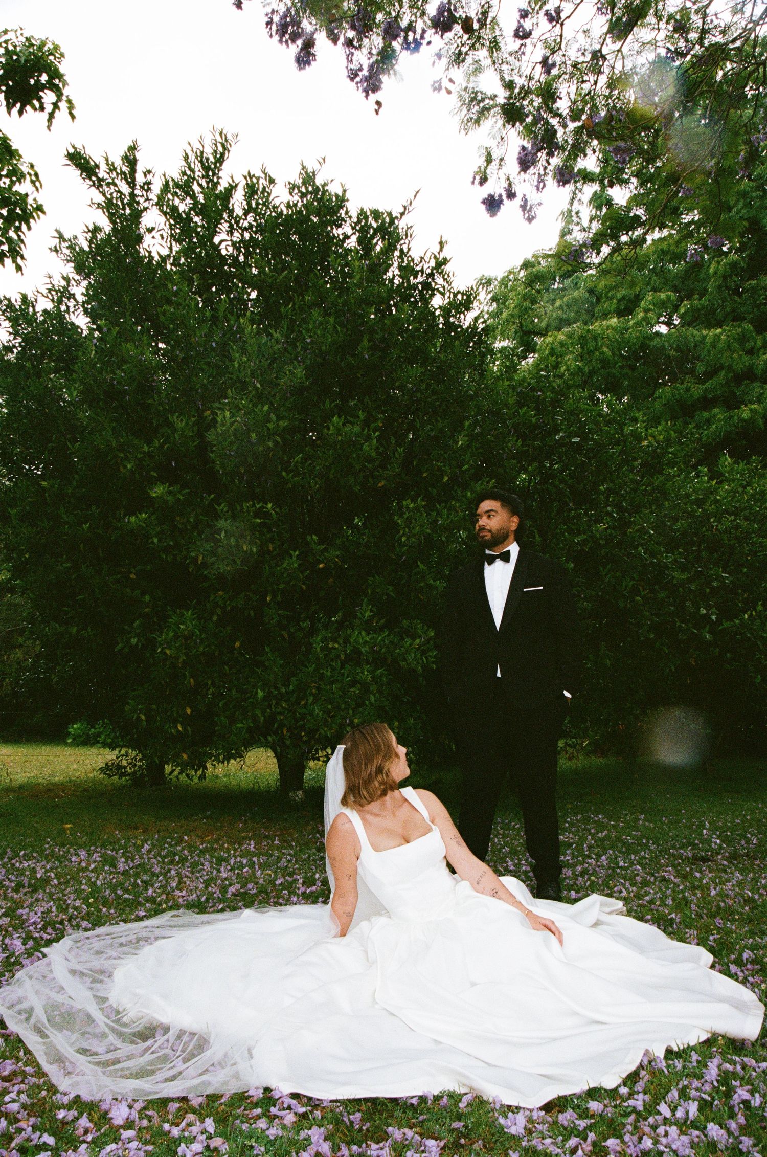 A bride in white sits gracefully on the ground while her groom stands among purple flowers.